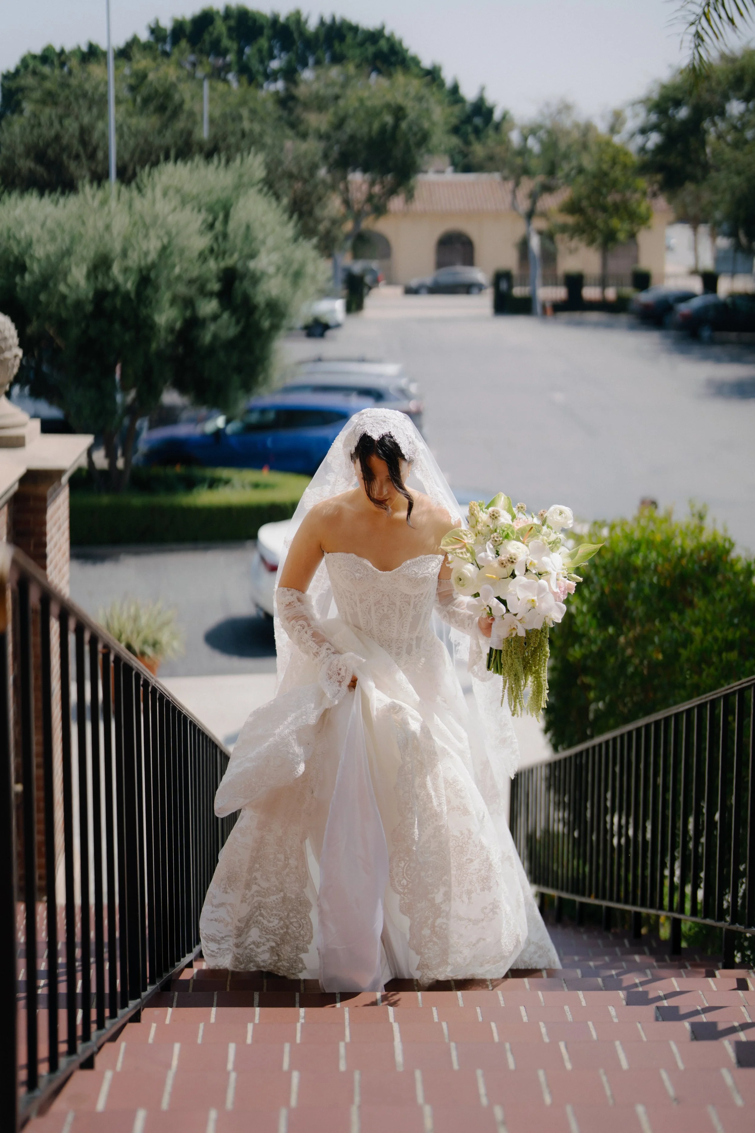 A bride in a white wedding gown and veil holding a bouquet of white flowers descending a brick staircase with a black railing outdoors on a sunny day.