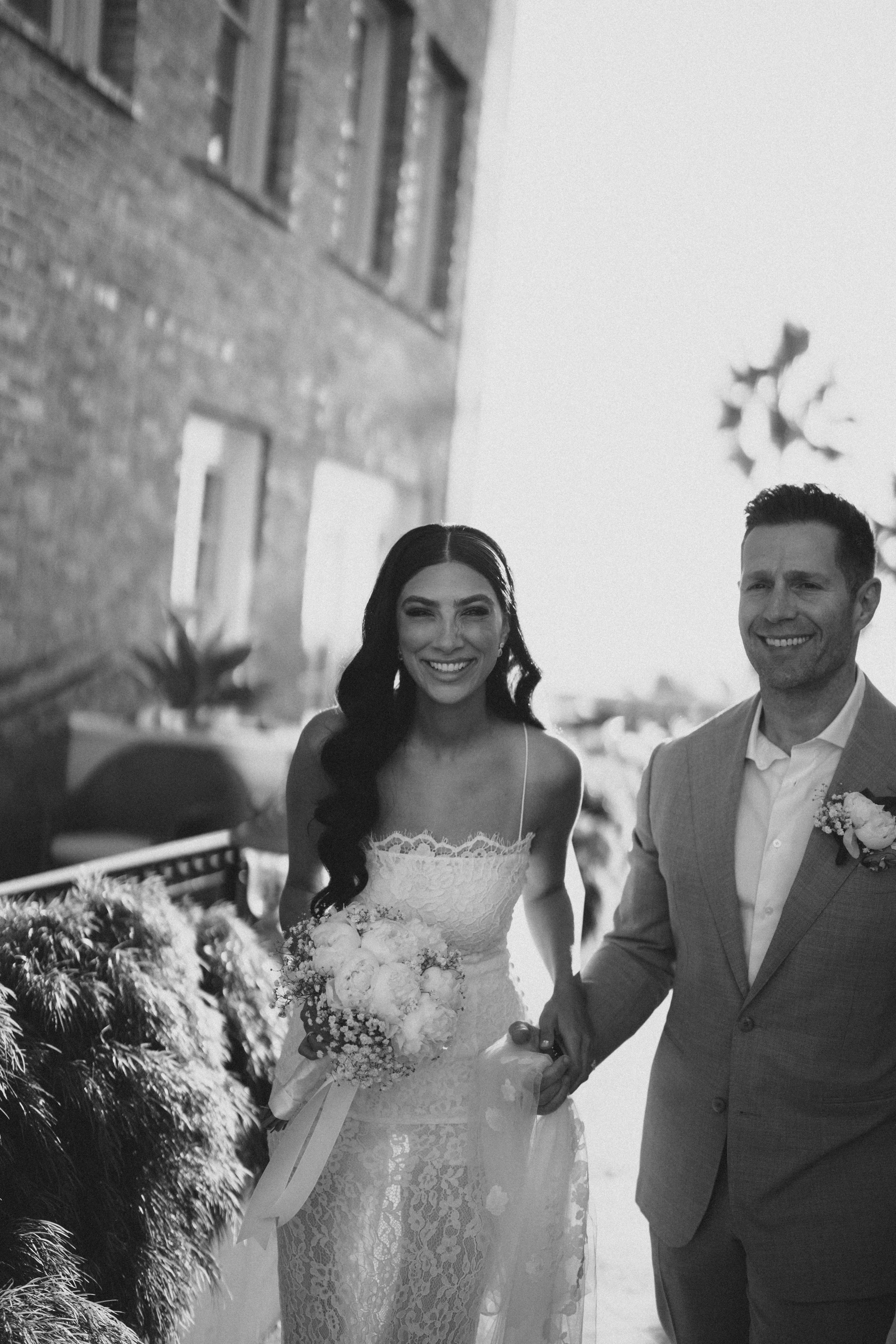 A bride and groom holding hands and smiling, outdoor wedding scene in black and white, with brick building and decorations in background.