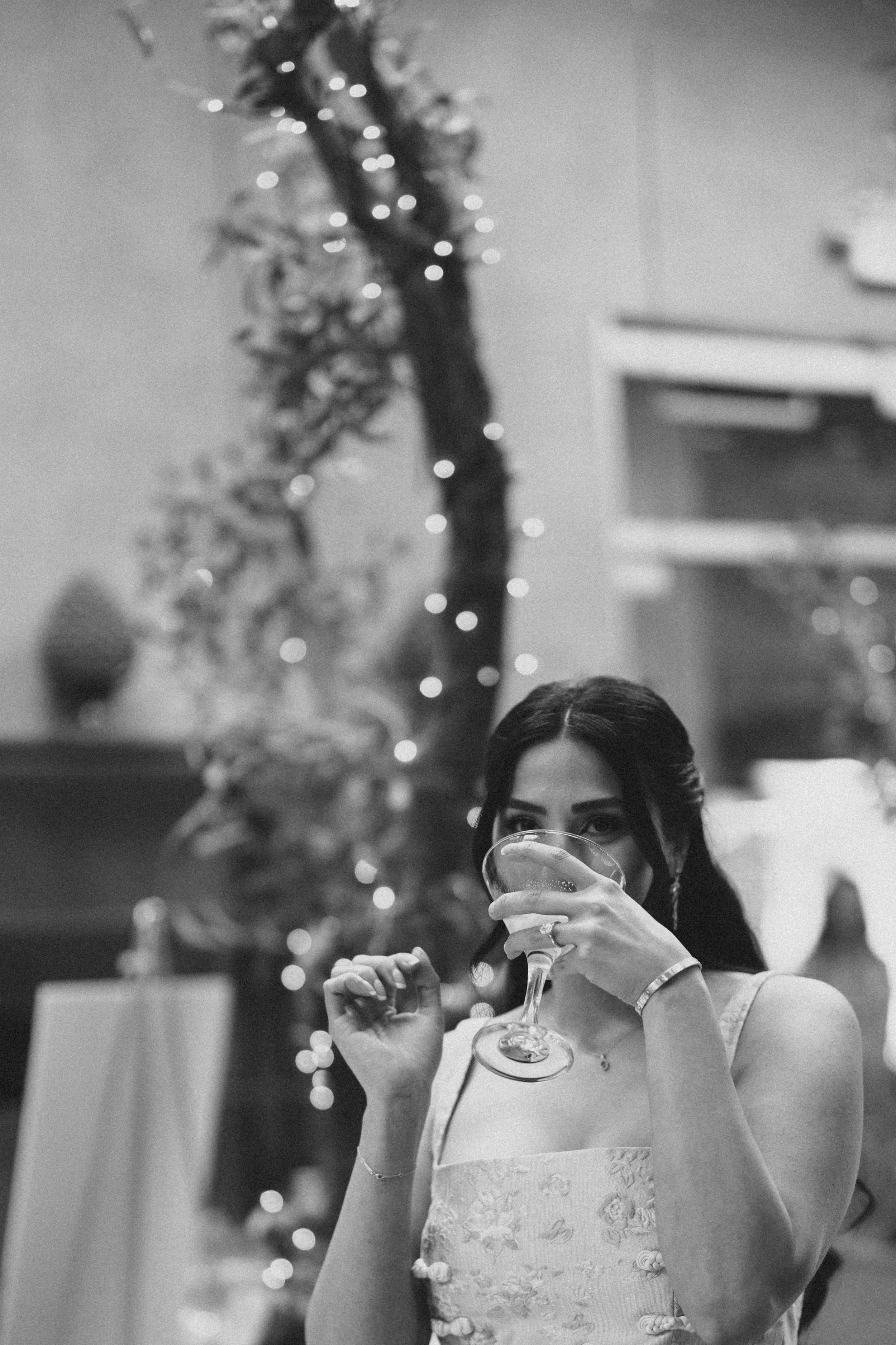 A woman with dark hair and earrings, wearing a sleeveless lace dress, holding a glass of champagne at a festive gathering with a decorated Christmas tree with lights in the background.