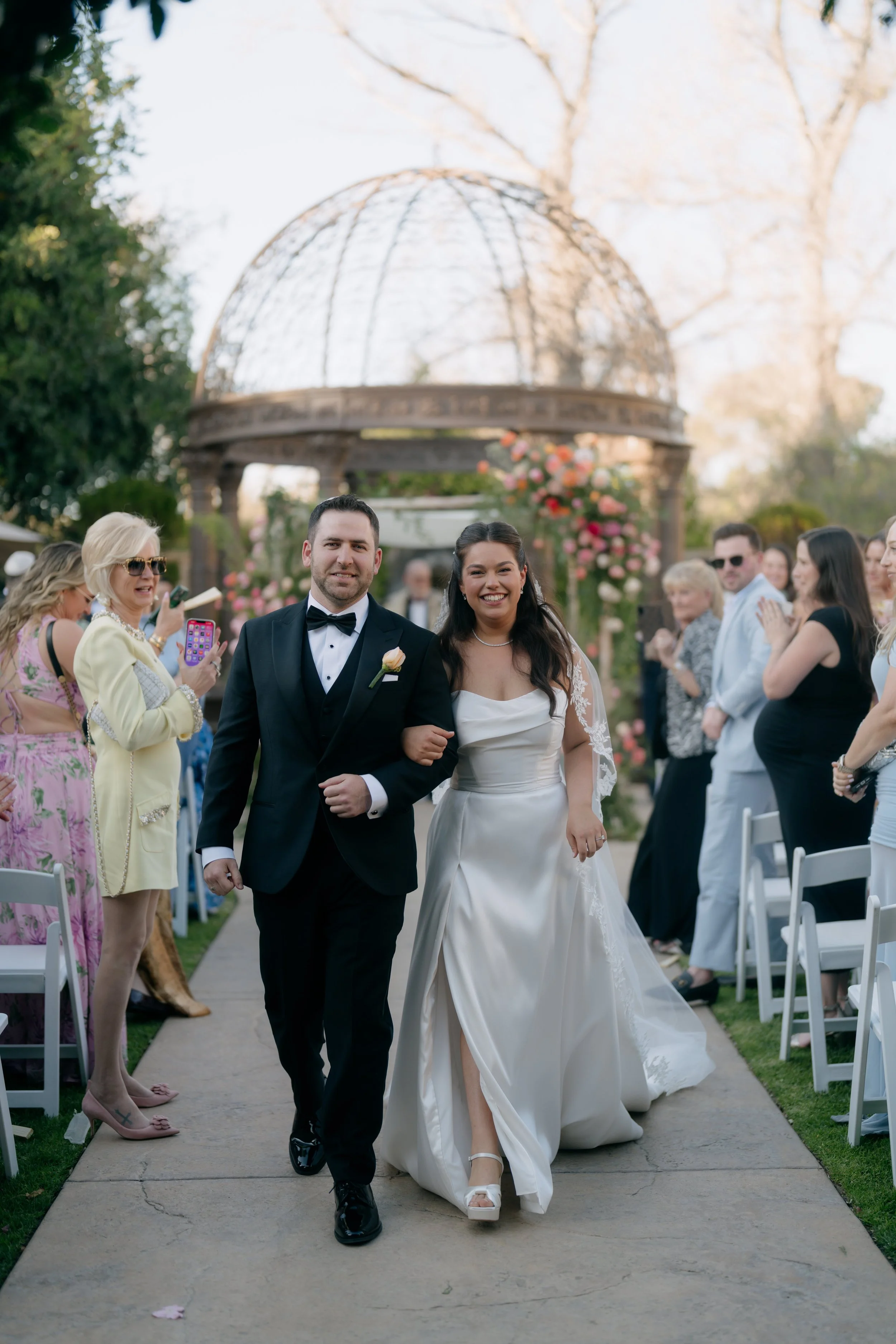 A bride and groom walking down an outdoor aisle during their wedding ceremony, smiling and holding hands, with guests on either side capturing the moment.