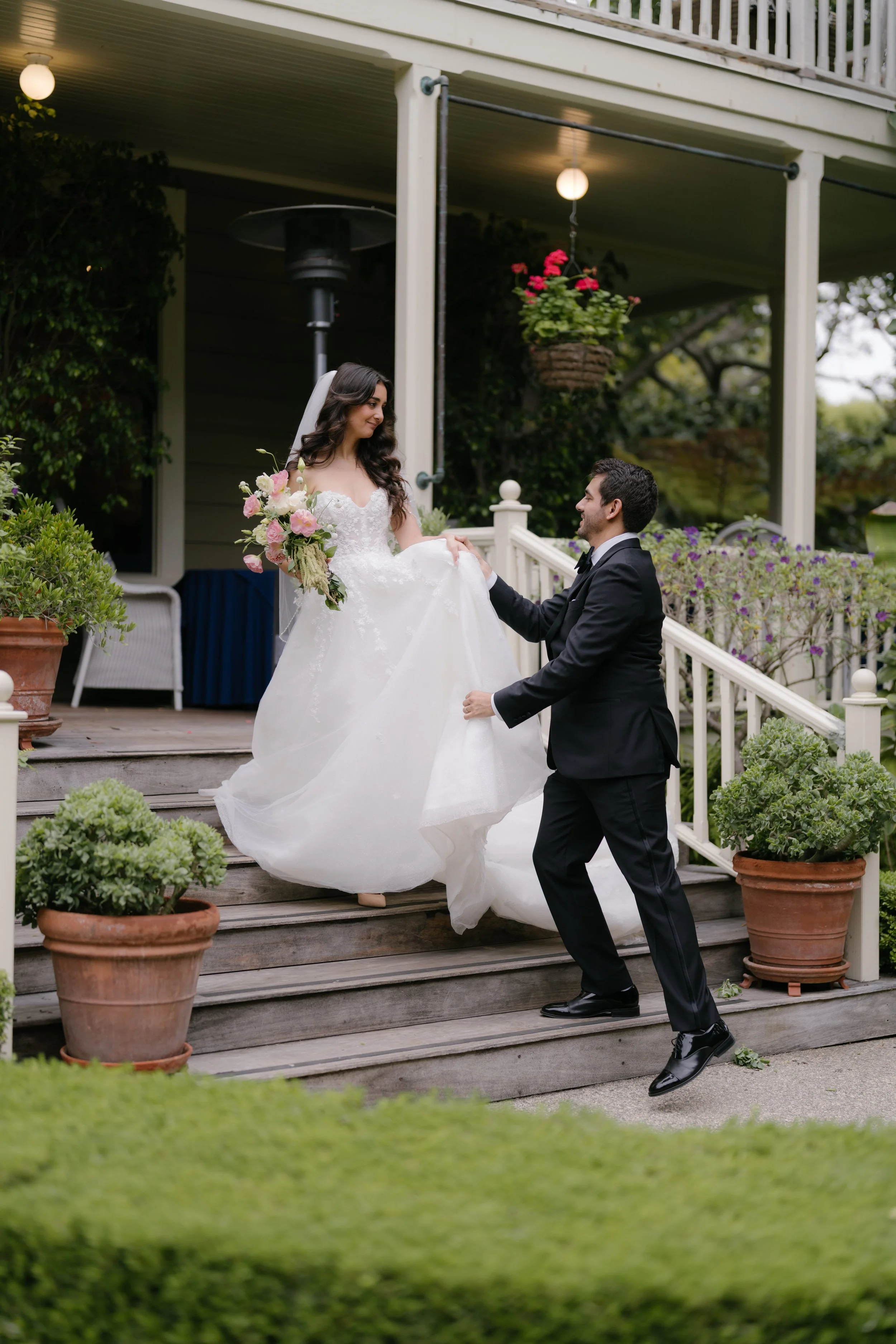 A bride and groom on a wooden porch surrounded by potted plants and hanging flowers. The groom is kneeling and holding the bride's hand, while the bride is holding a bouquet of pink and white flowers and smiling at him.