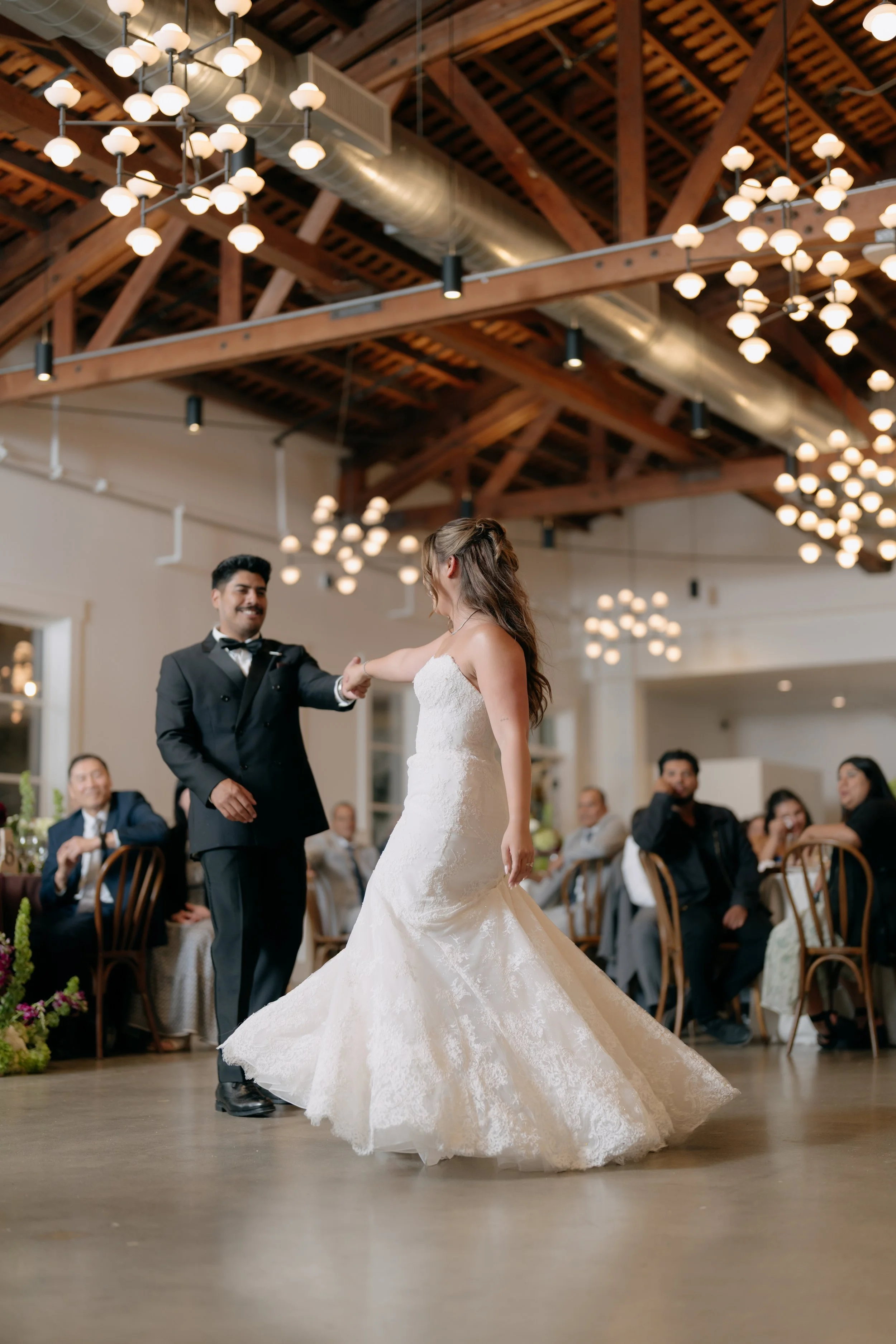 A bride and groom are dancing during their wedding reception, with guests seated around them watching and smiling. The venue has a high ceiling with wooden beams and modern lighting.