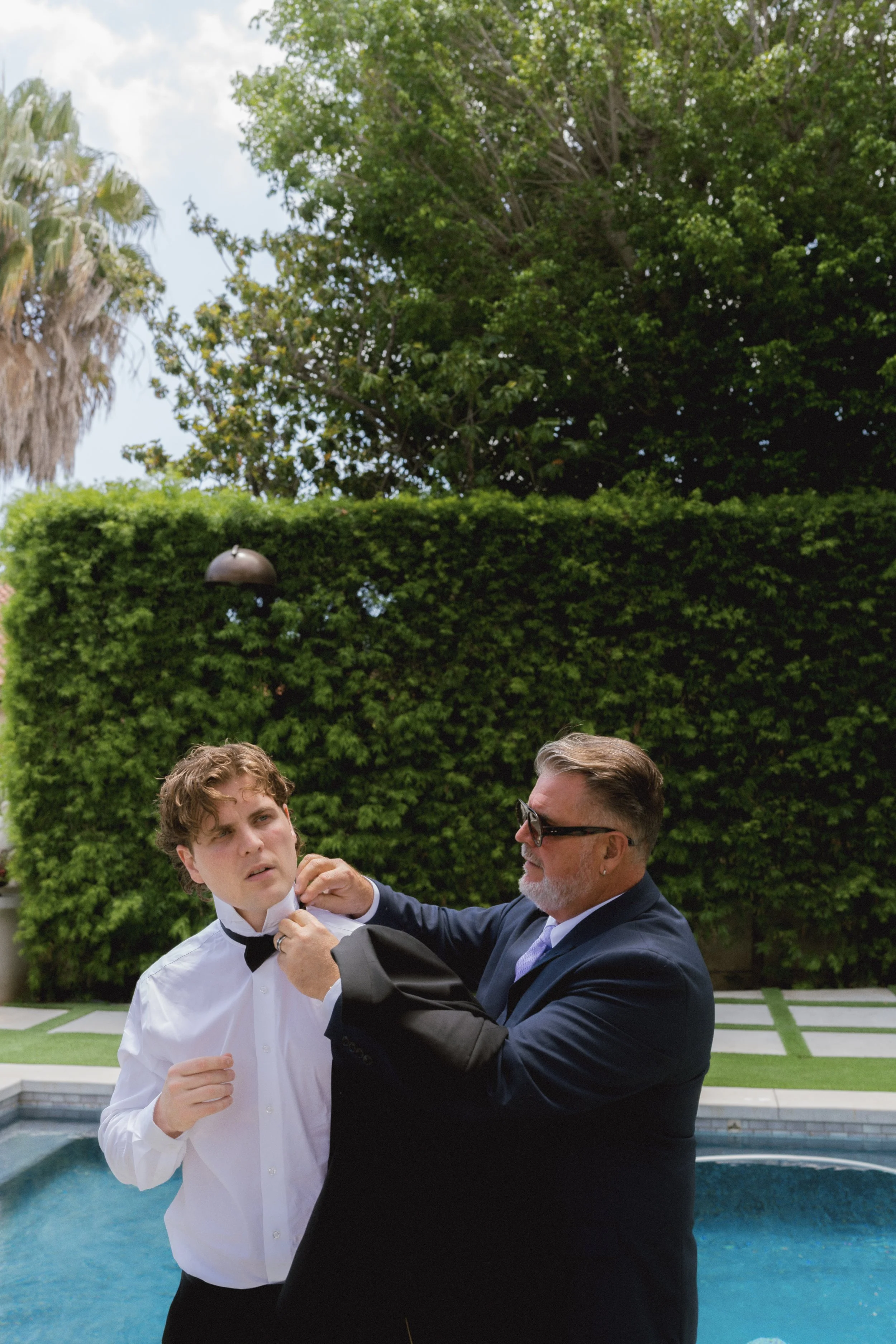 A young man in a white dress shirt and black bowtie being adjusted by an older man in a suit and sunglasses outdoors near a pool with a manicured hedge and trees in the background.