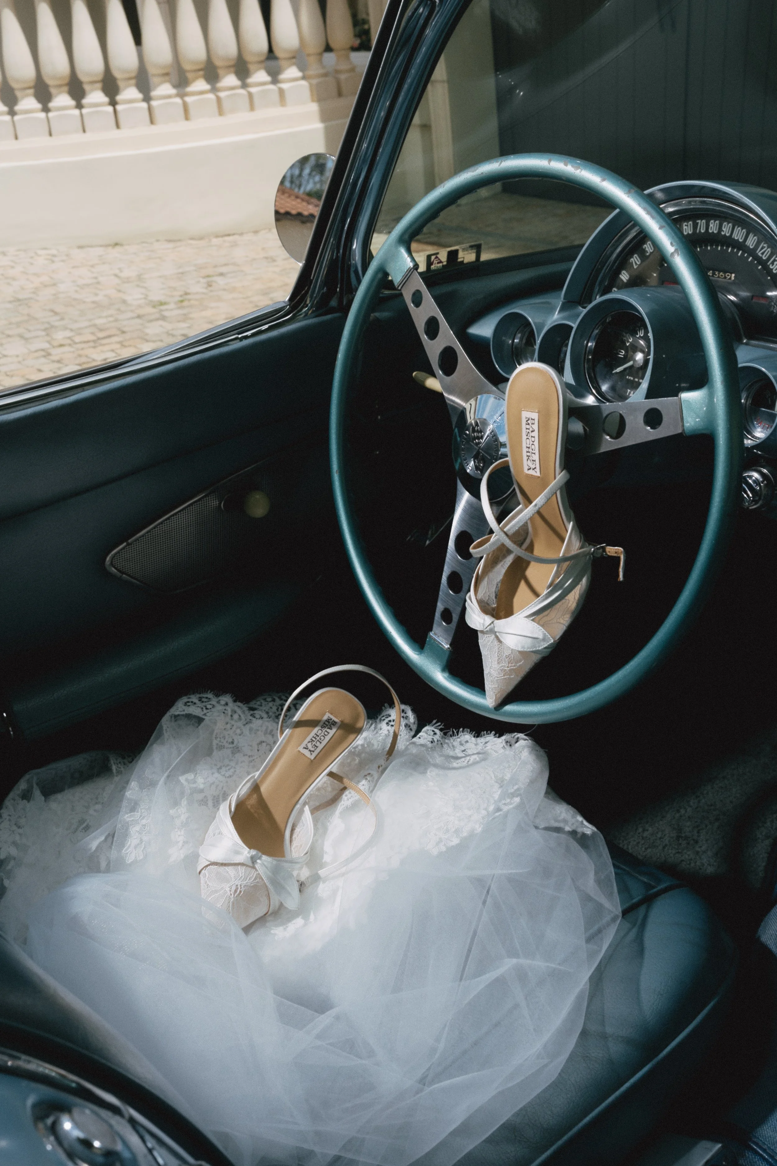 The interior of a vintage car with a teal steering wheel, wedding shoes resting on a white tulle cloth, and a glimpse of the car’s speedometer and side mirror.