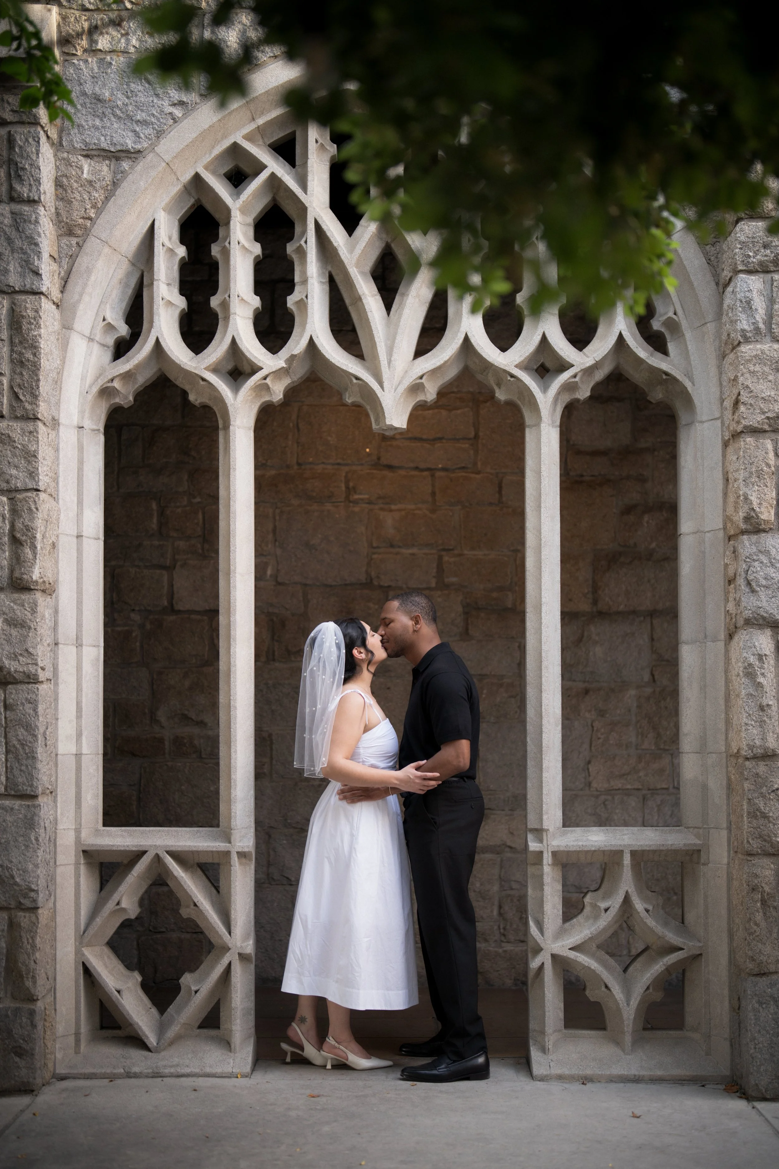 A bride and groom kissing under a decorative stone arch, with the bride in a white dress and veil, and the groom in a black shirt and pants.