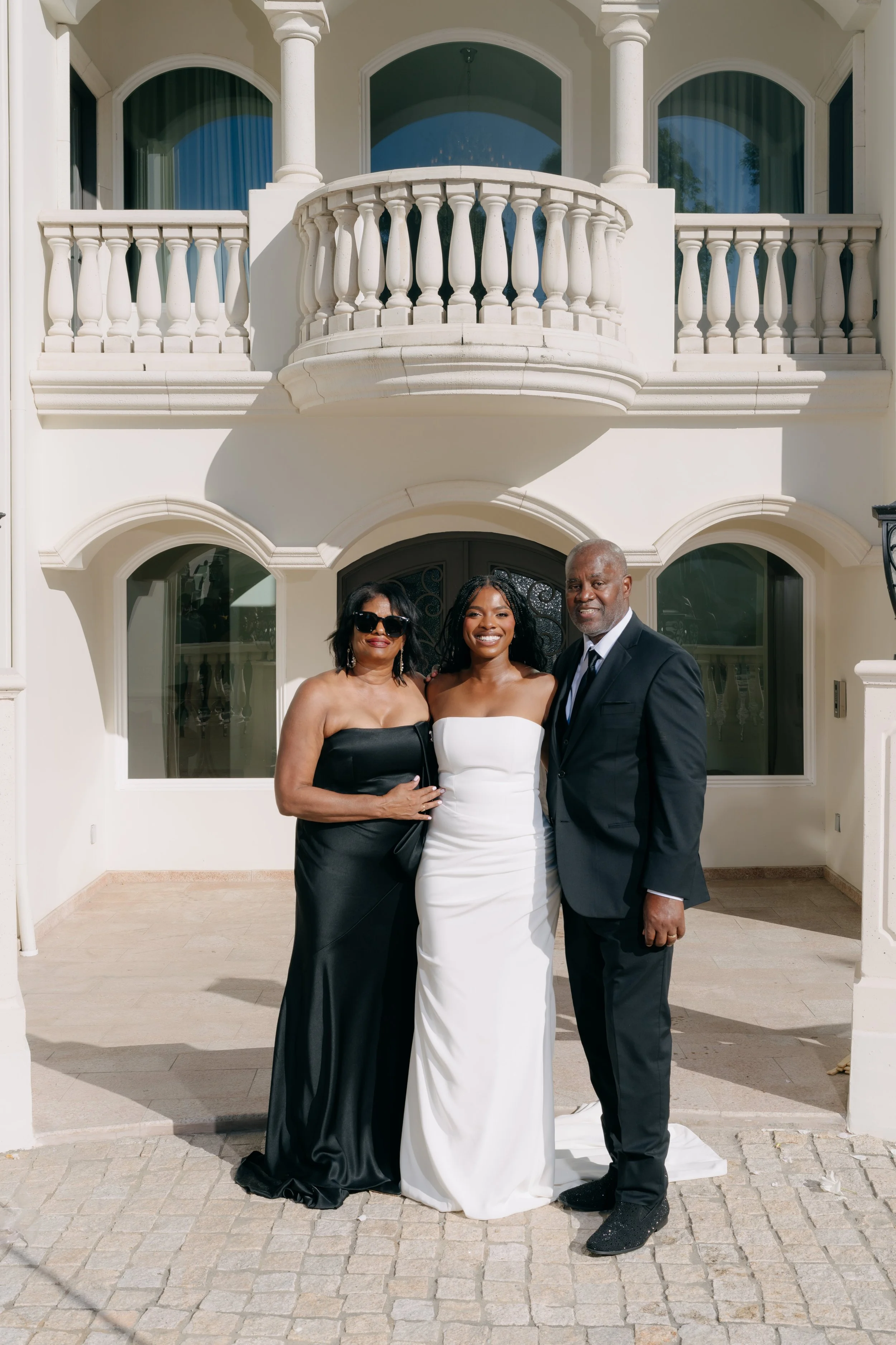 Three people standing outdoors in front of a large, ornate white house with a rounded balcony and columns, dressed formally for a wedding or special occasion.