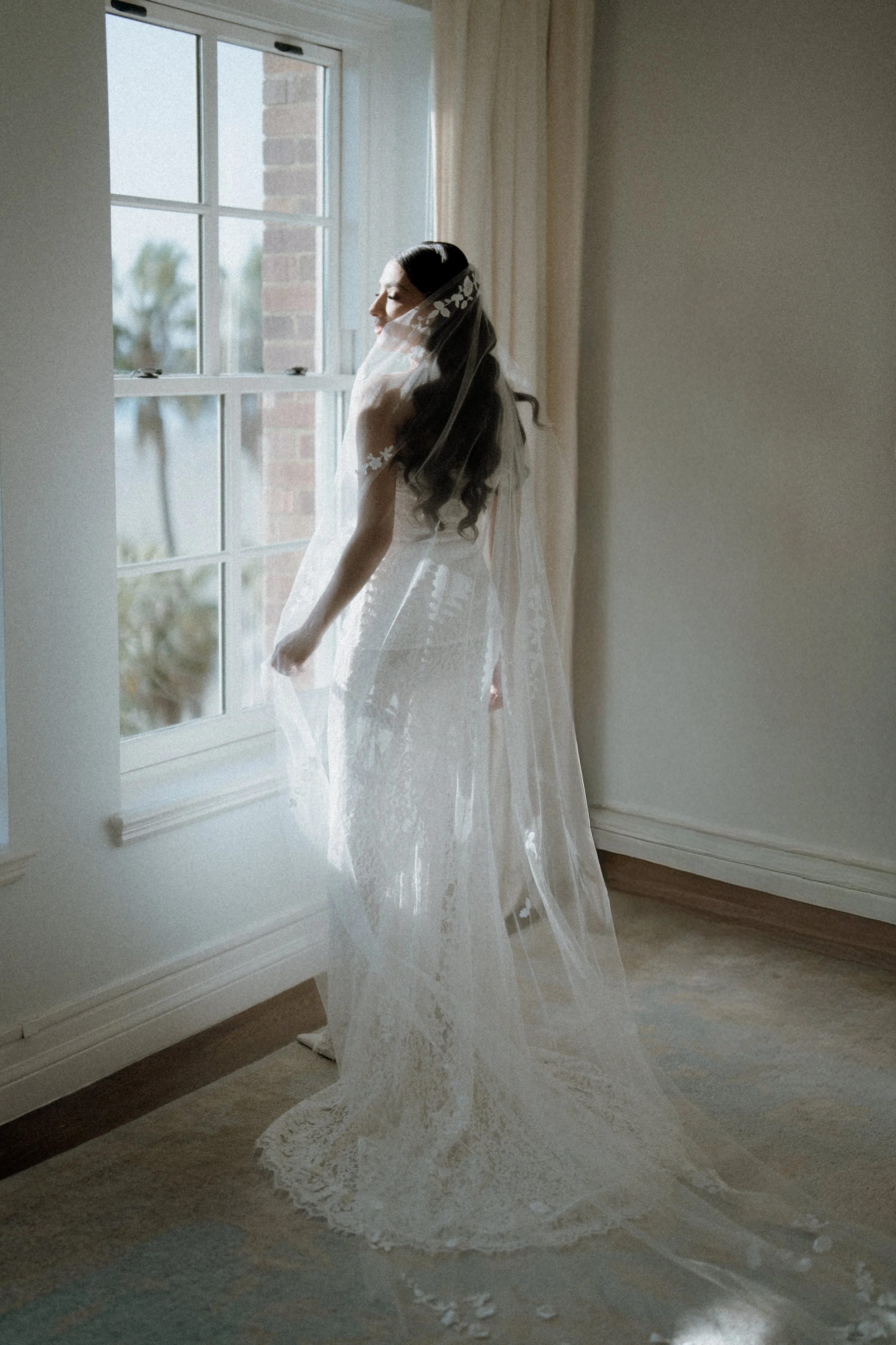Bride in a white wedding gown and veil standing by a window with sunlight streaming in, inside a room with light-colored walls and wooden flooring.
