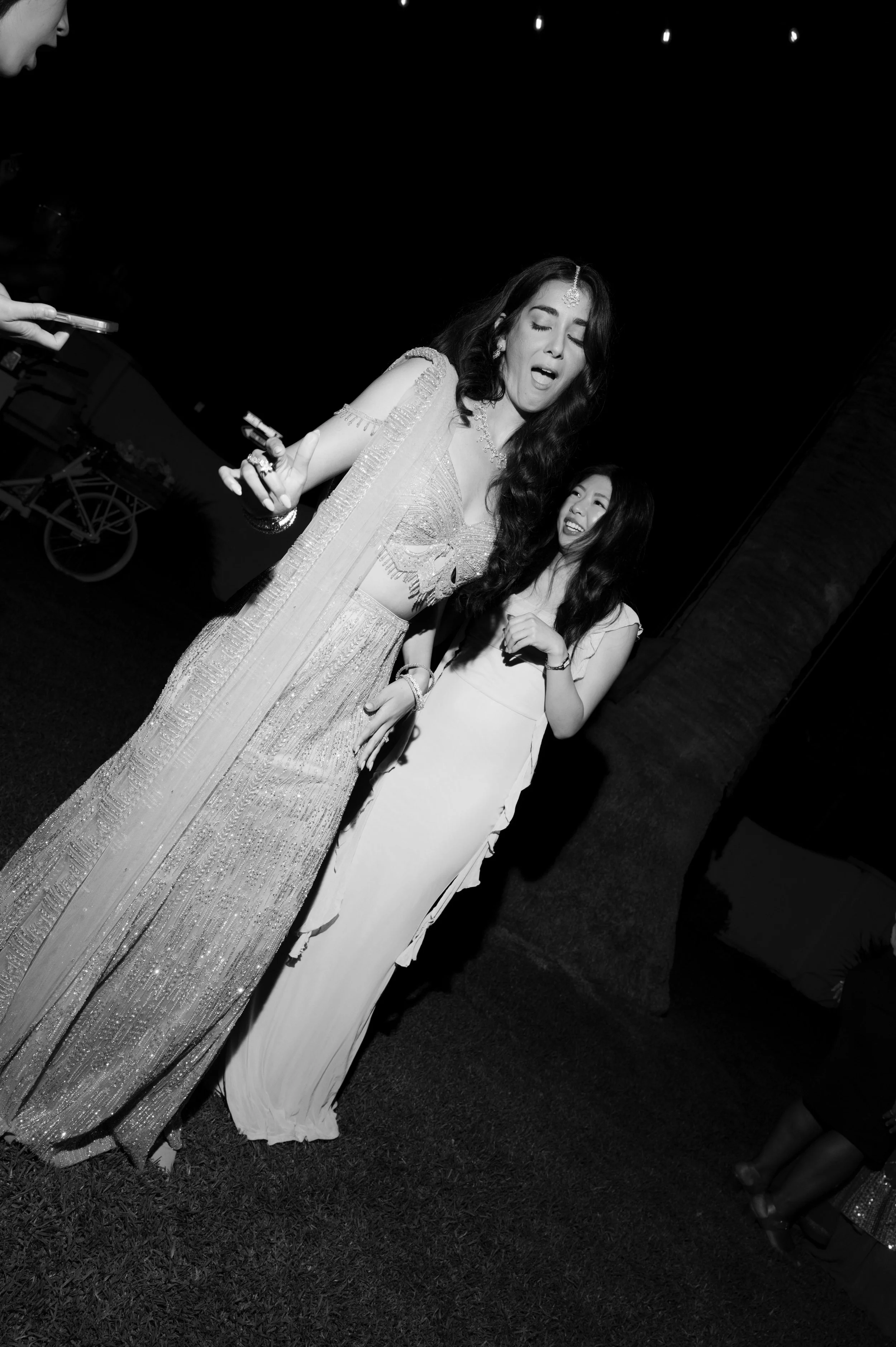 A woman in traditional Indian attire singing or speaking at night, with another woman smiling nearby, during an outdoor event with string lights overhead.