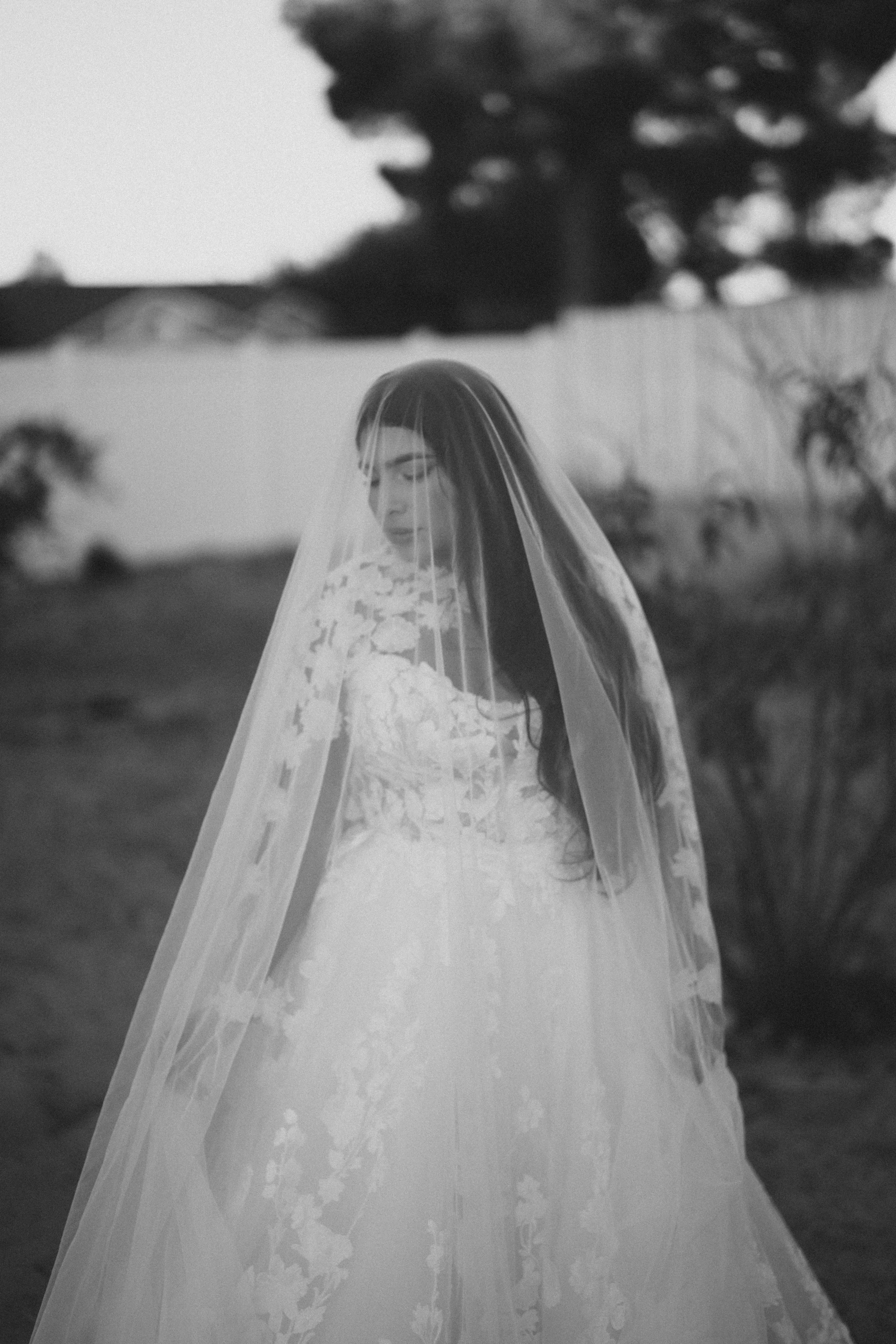 Black and white photo of a woman in a wedding dress with a veil, standing outdoors in front of trees and a fence, looking down with a serene expression.