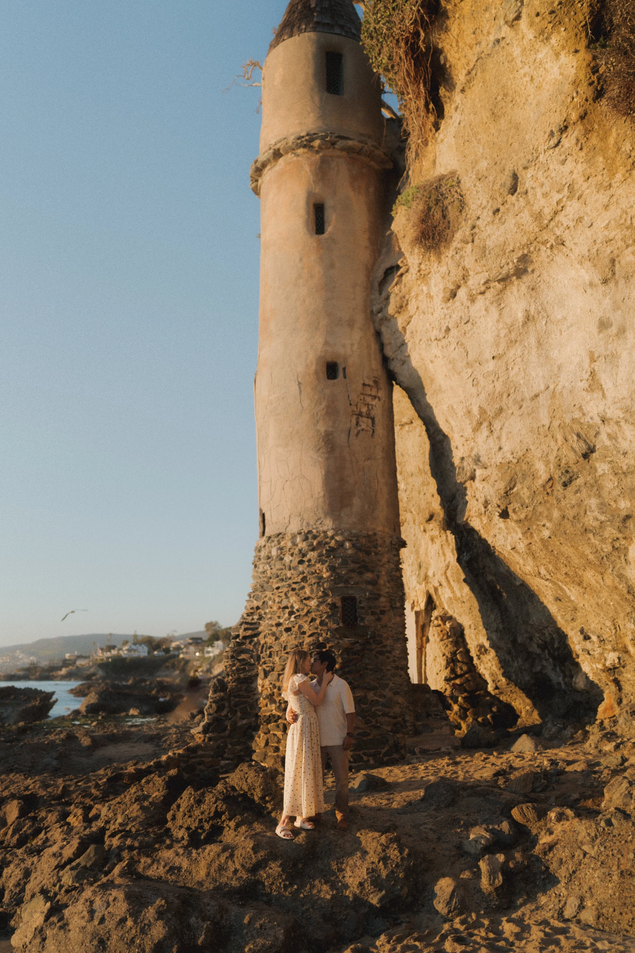 A couple embracing and kissing on a rocky beach at sunset, with a tall, circular stone tower and castle ruins in the background, overlooking the ocean and a nearby town.