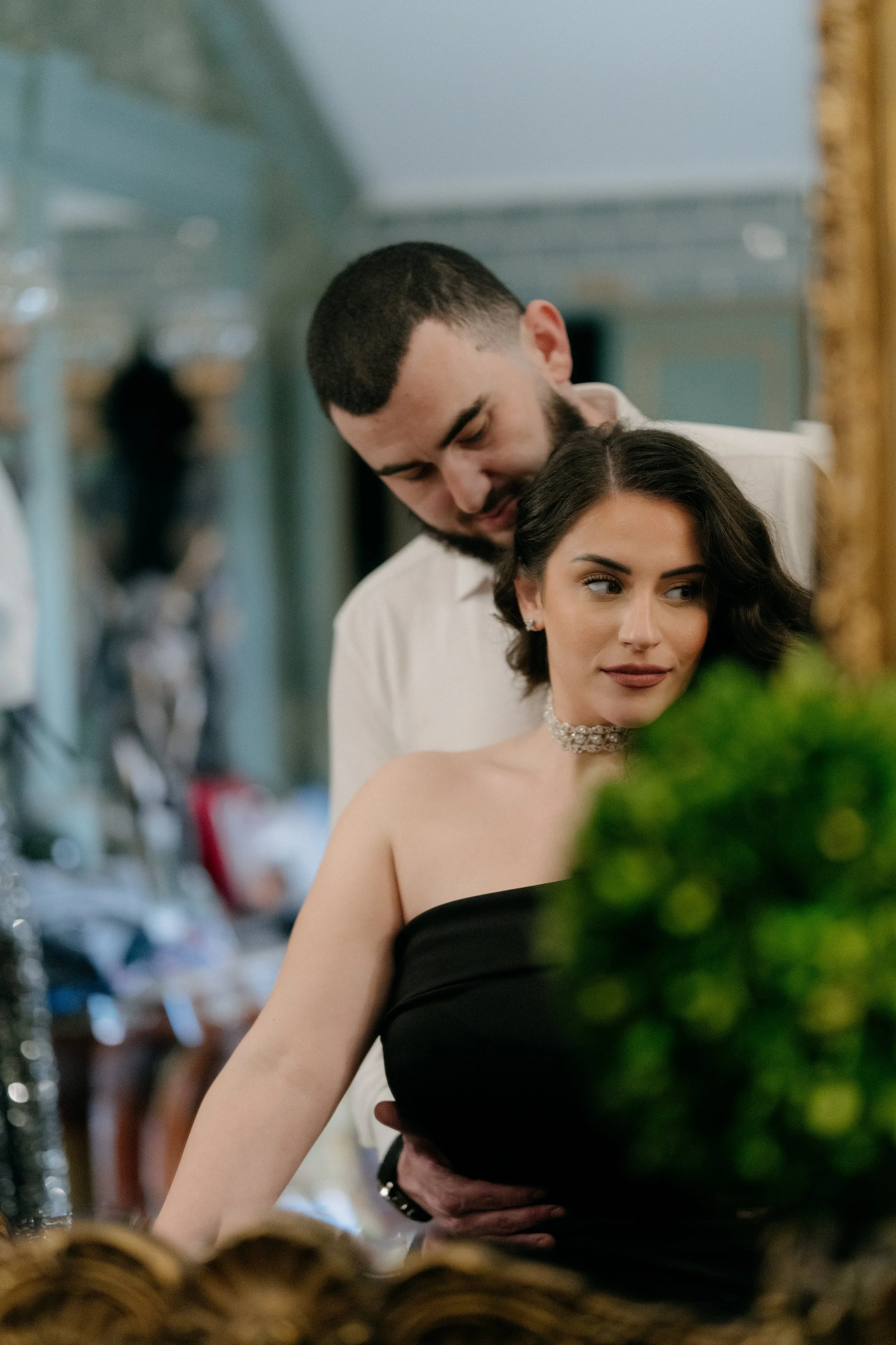 A man and woman dressed formally, possibly at a wedding or event, standing close together with the man leaning over the woman. The woman has dark hair and wears a choker and strapless dress, while the man has short dark hair and a beard, wearing a wh