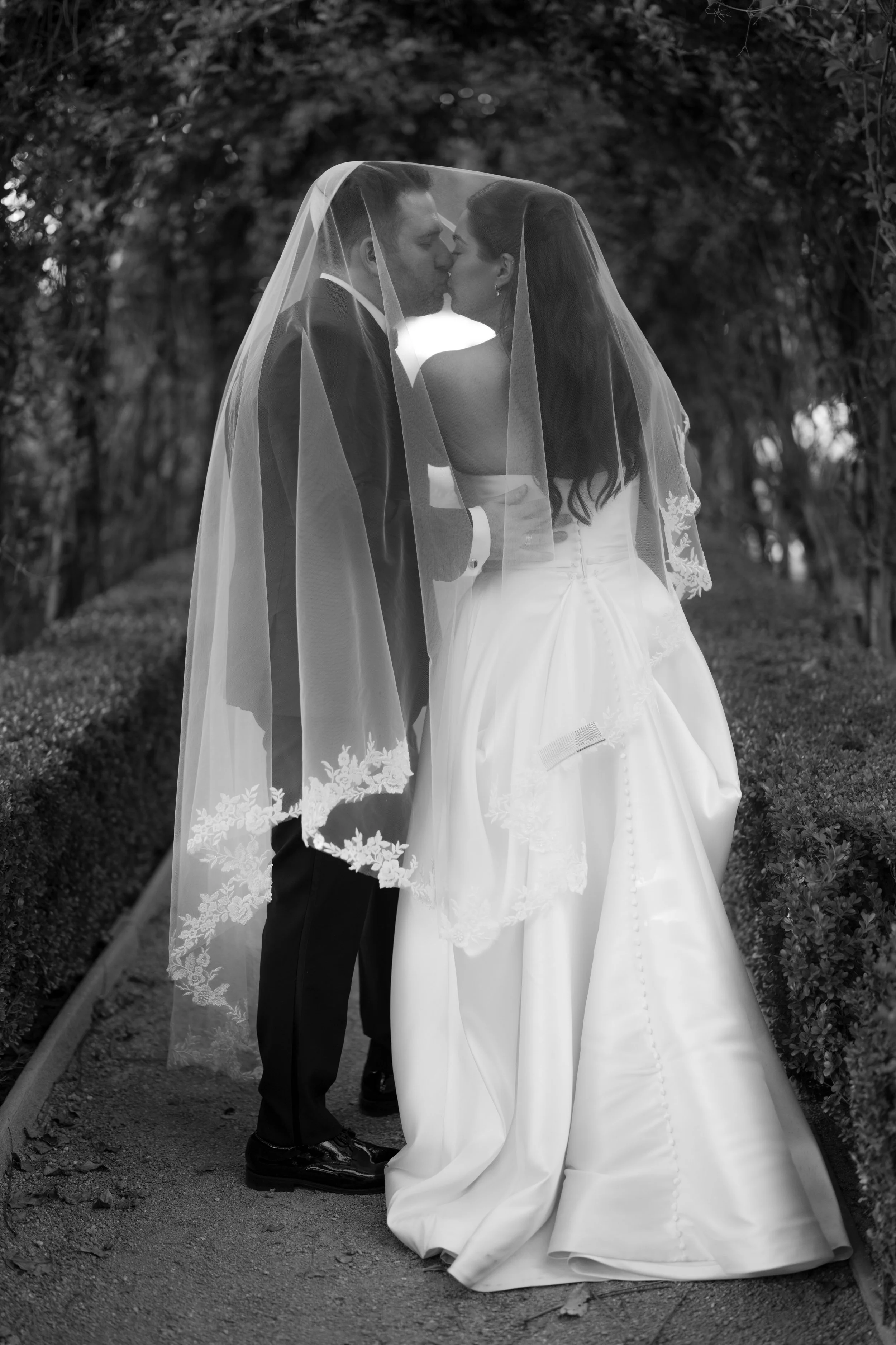 A black and white photo of a bride and groom kissing underneath a veil on their wedding day outdoors, surrounded by trees.