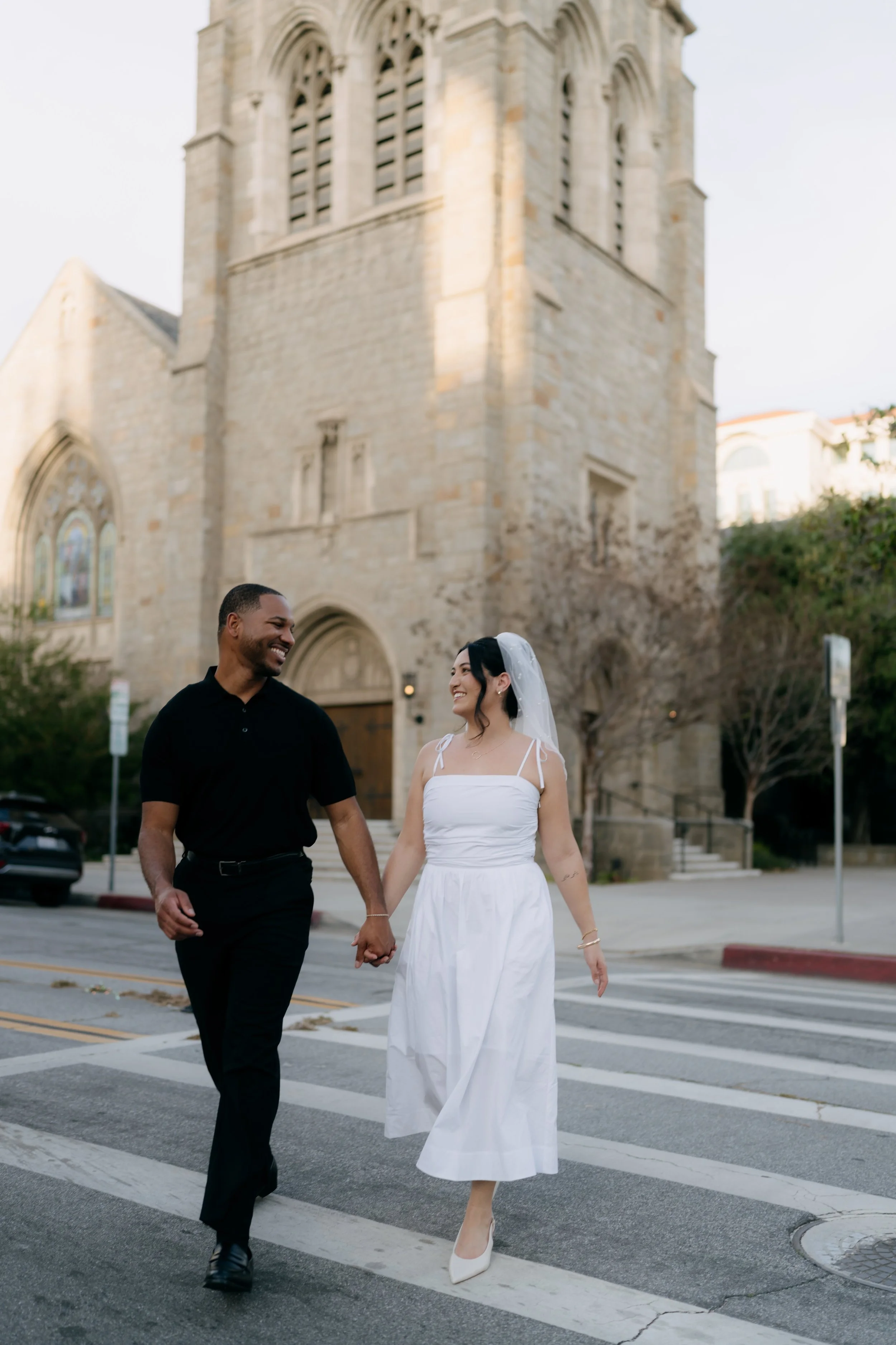 A happy interracial couple walking hand in hand across the street in front of a historic church, with the woman wearing a white dress and veil, and the man in a black shirt and pants, in an urban setting with trees and parked cars.