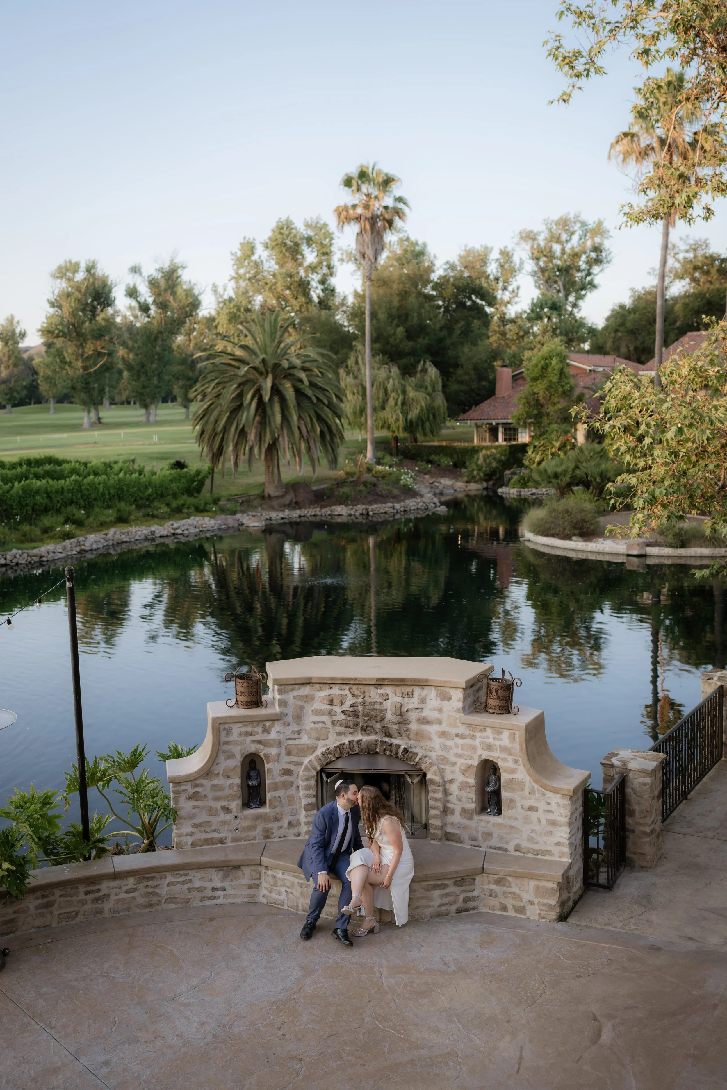 A couple dressed in wedding attire standing on a stone terrace beside a small water fountain, with a scenic background of a pond, trees, and a house. The groom is in a gray suit and the bride is in a white wedding dress.