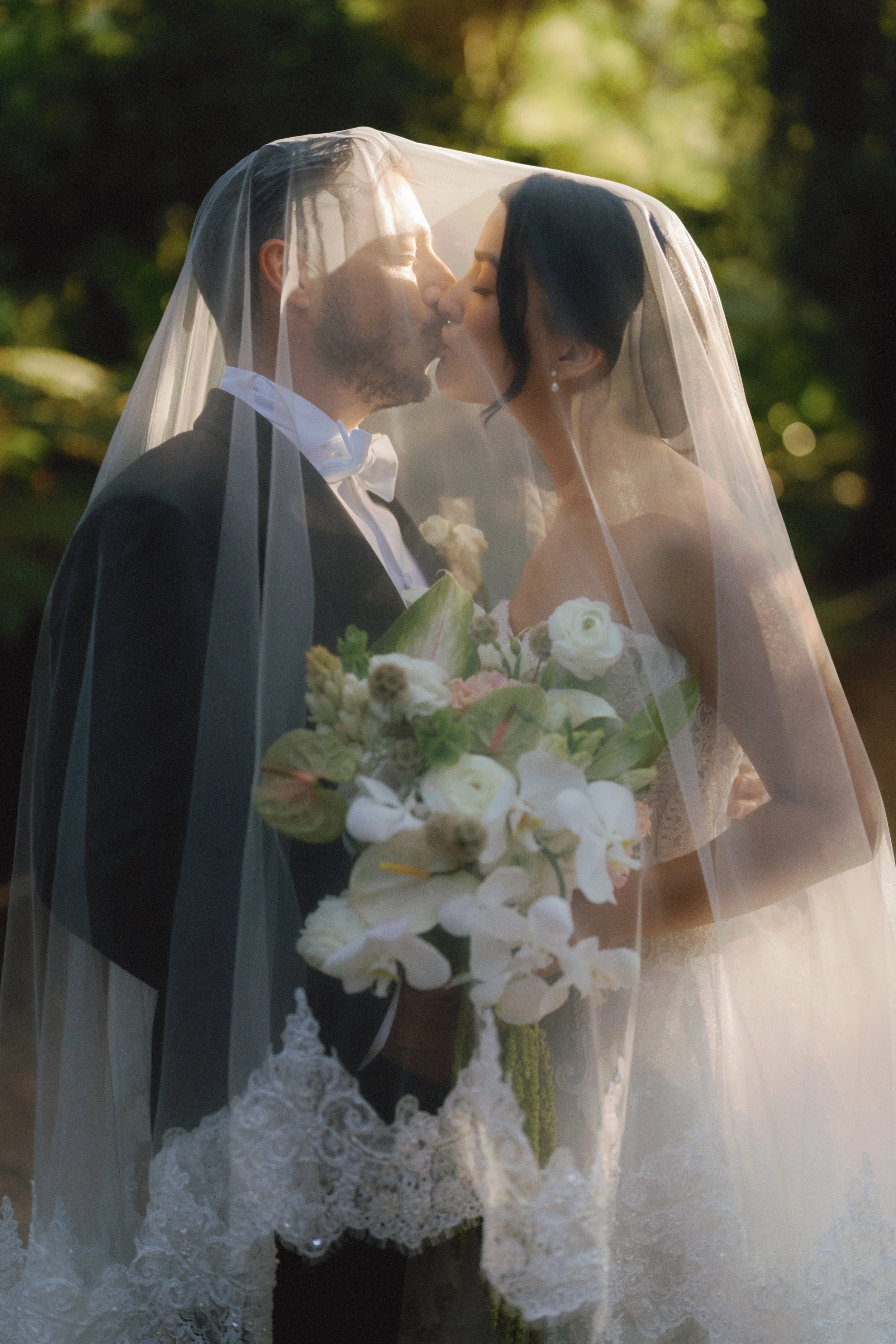 A bride and groom sharing a kiss under a veil, outdoors, with the bride holding a bouquet of white and pink flowers, in a romantic wedding setting.