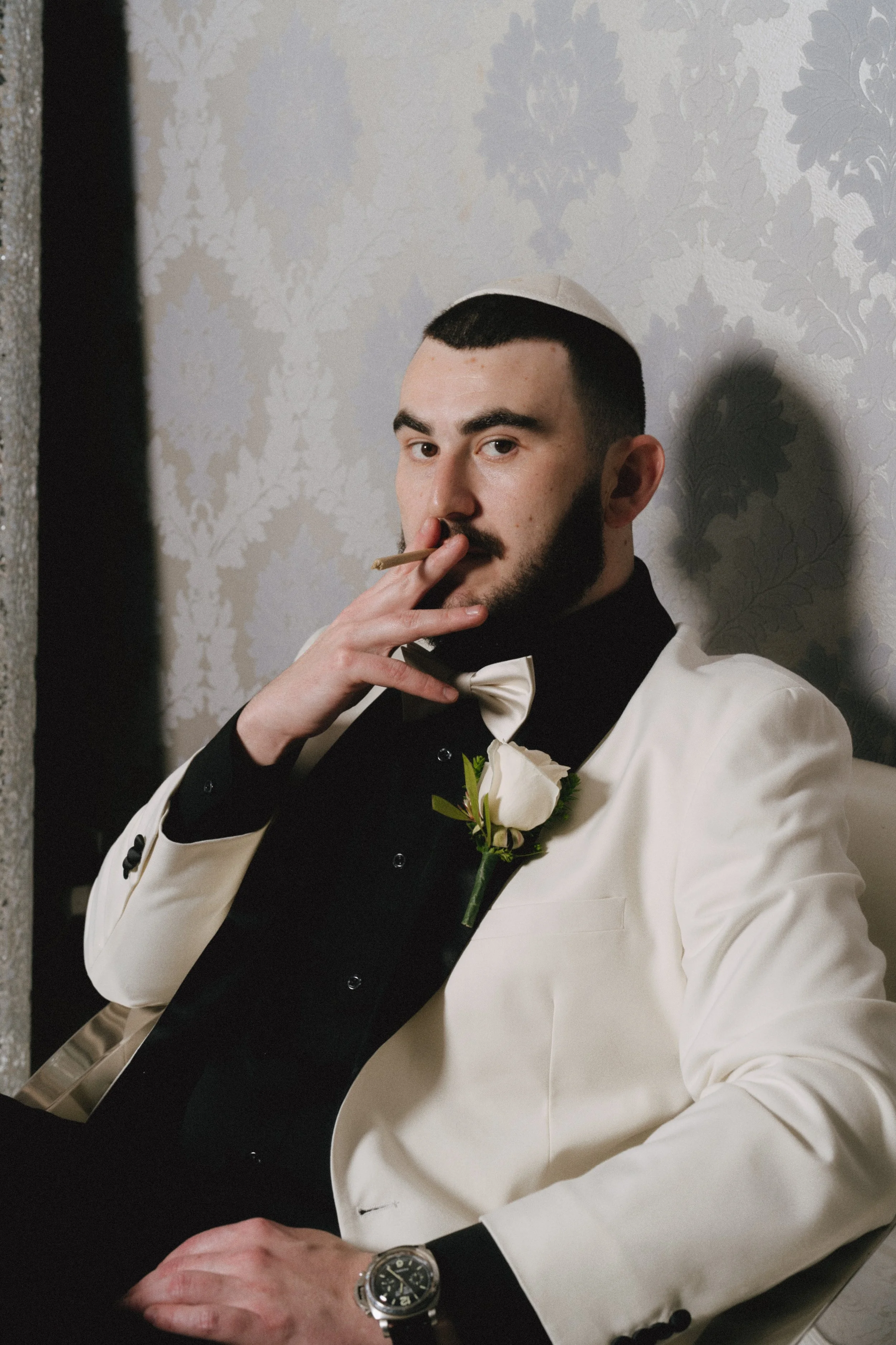 A man dressed in a tuxedo with a white jacket and black waistcoat, wearing a bow tie and a boutonniere on his lapel, is smoking a cigar. He is sitting with his hand resting on a table, wearing a wristwatch. The background features a gray wallpaper wi