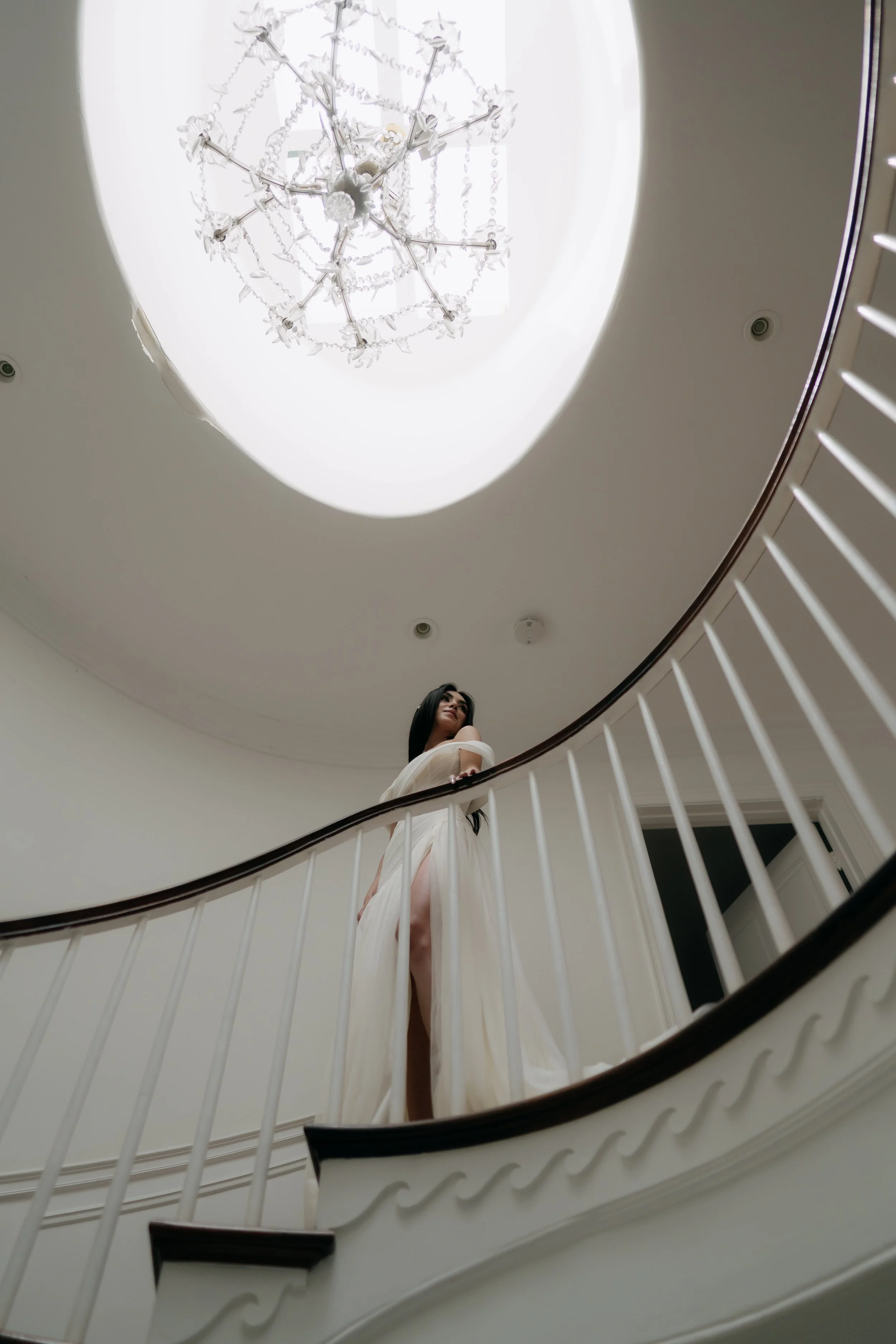 A woman wearing a white dress standing on a staircase, with a spiral staircase railing in the foreground, viewed from below.