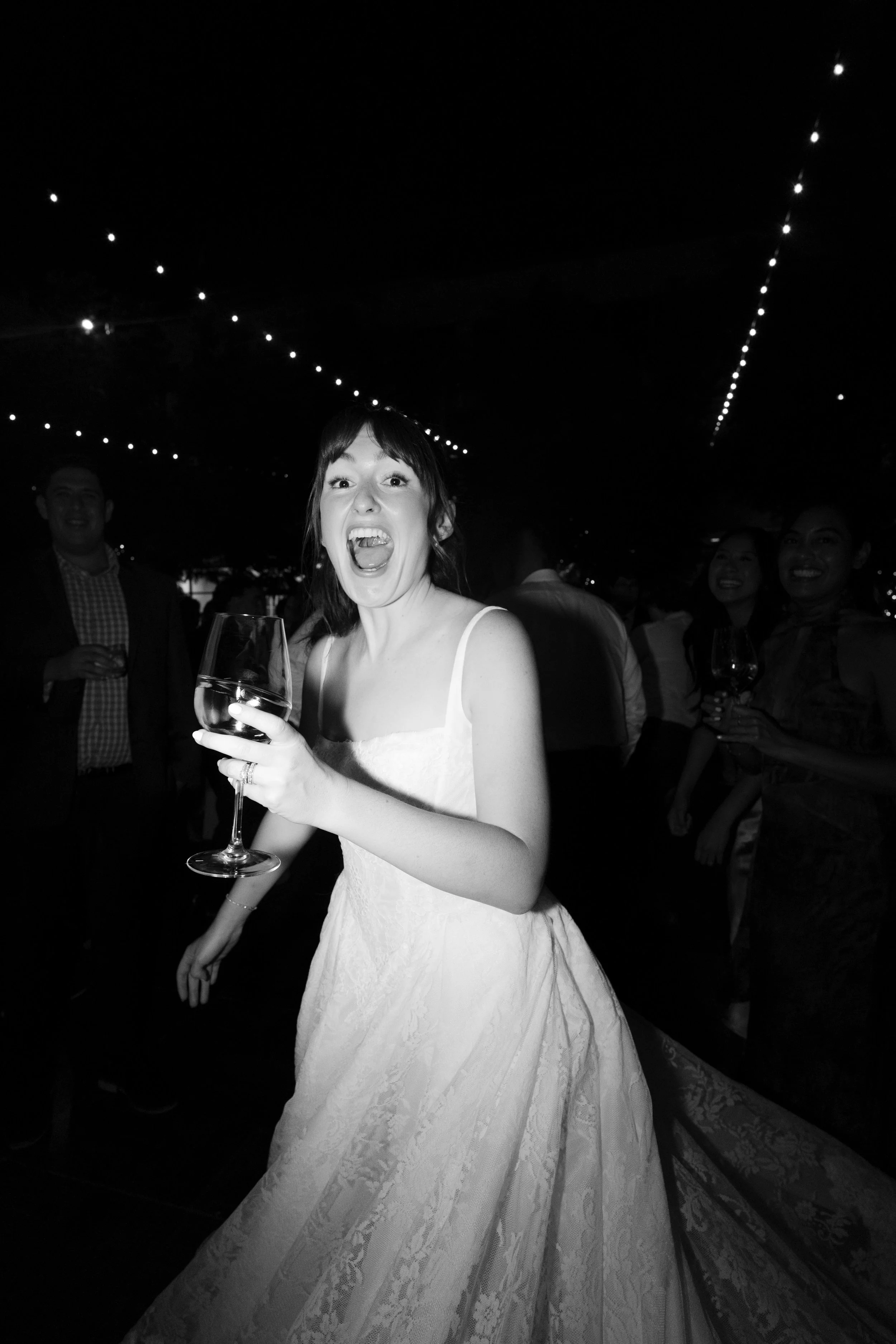 Black and white photo of a woman in a white dress at a party, holding a wine glass and smiling with her mouth open, surrounded by other people with drinks, under string lights.