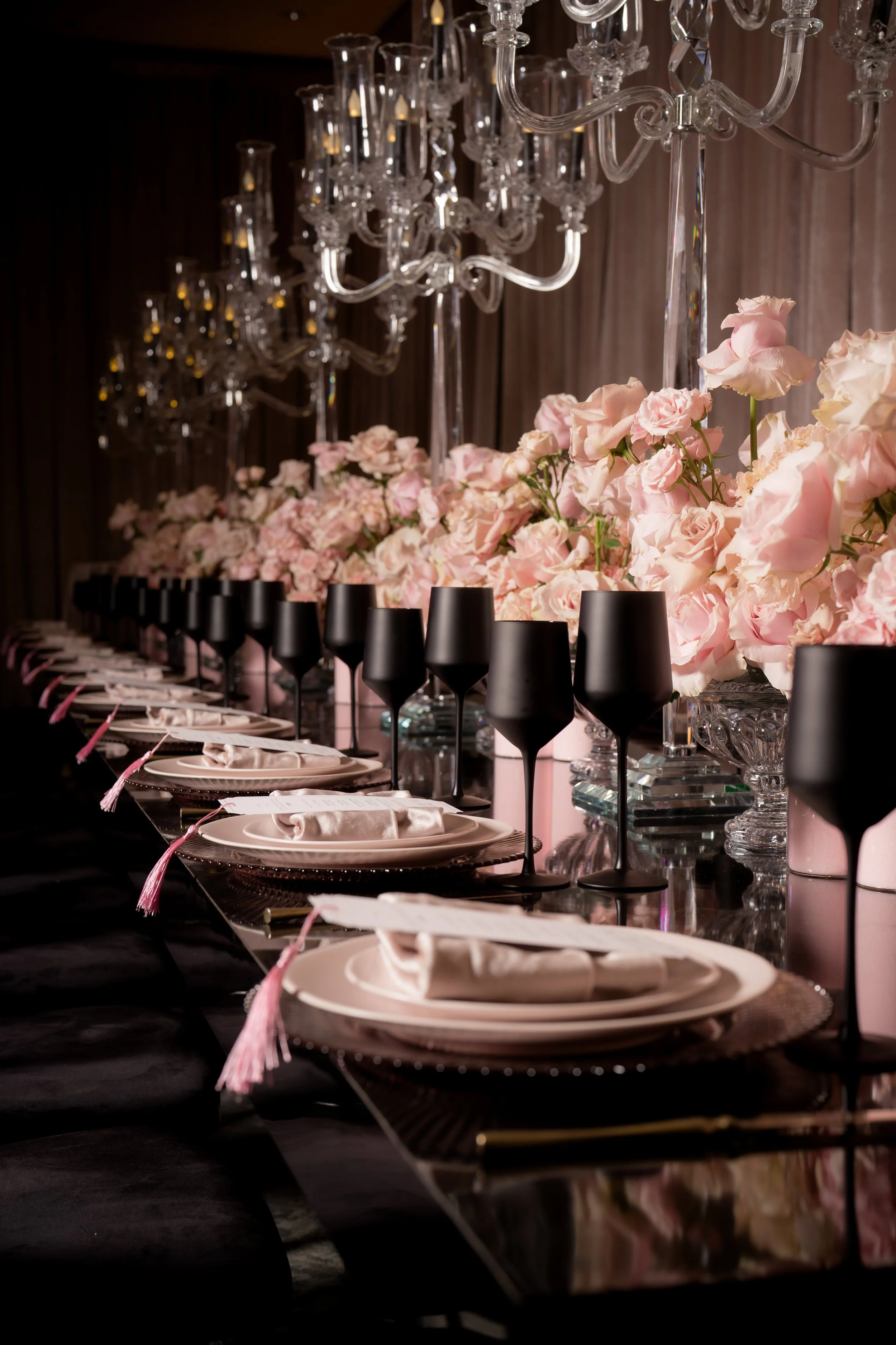 A dinner table decorated with pink roses, black wine glasses, and white plates with napkins, with large crystal chandeliers hanging above, in a dimly lit setting.