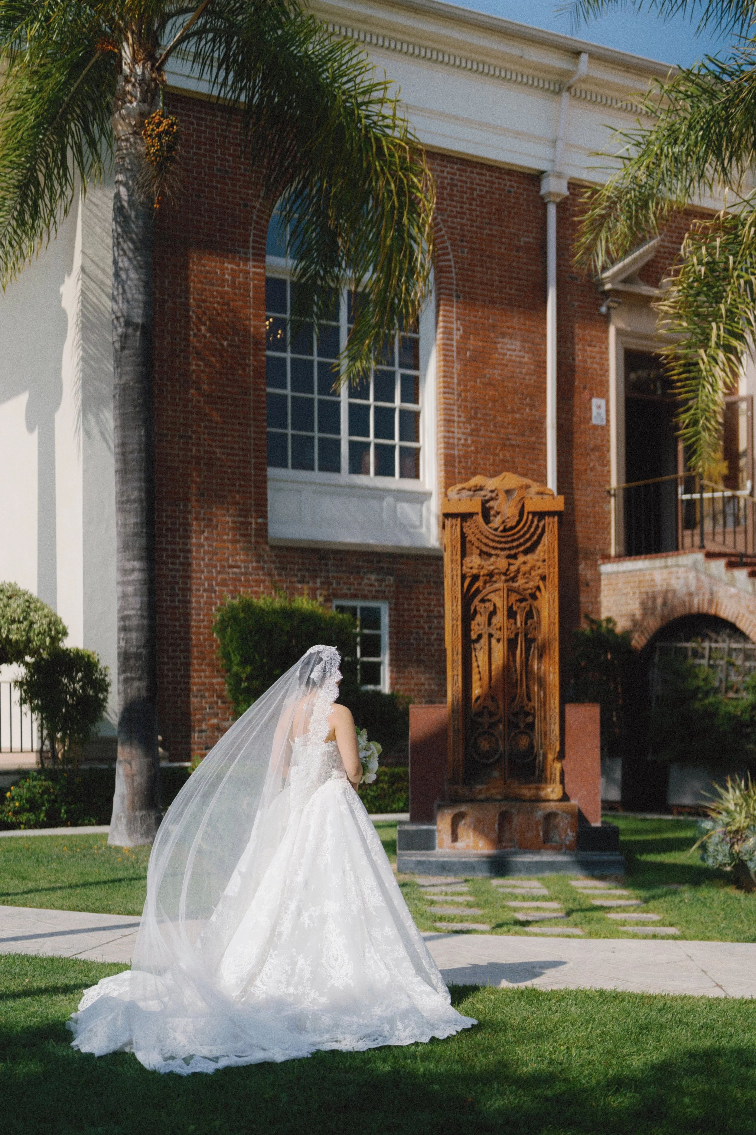 A bride in a white wedding gown and veil stands outdoors near a historical wooden sculpture, with a brick building and palm trees in the background.