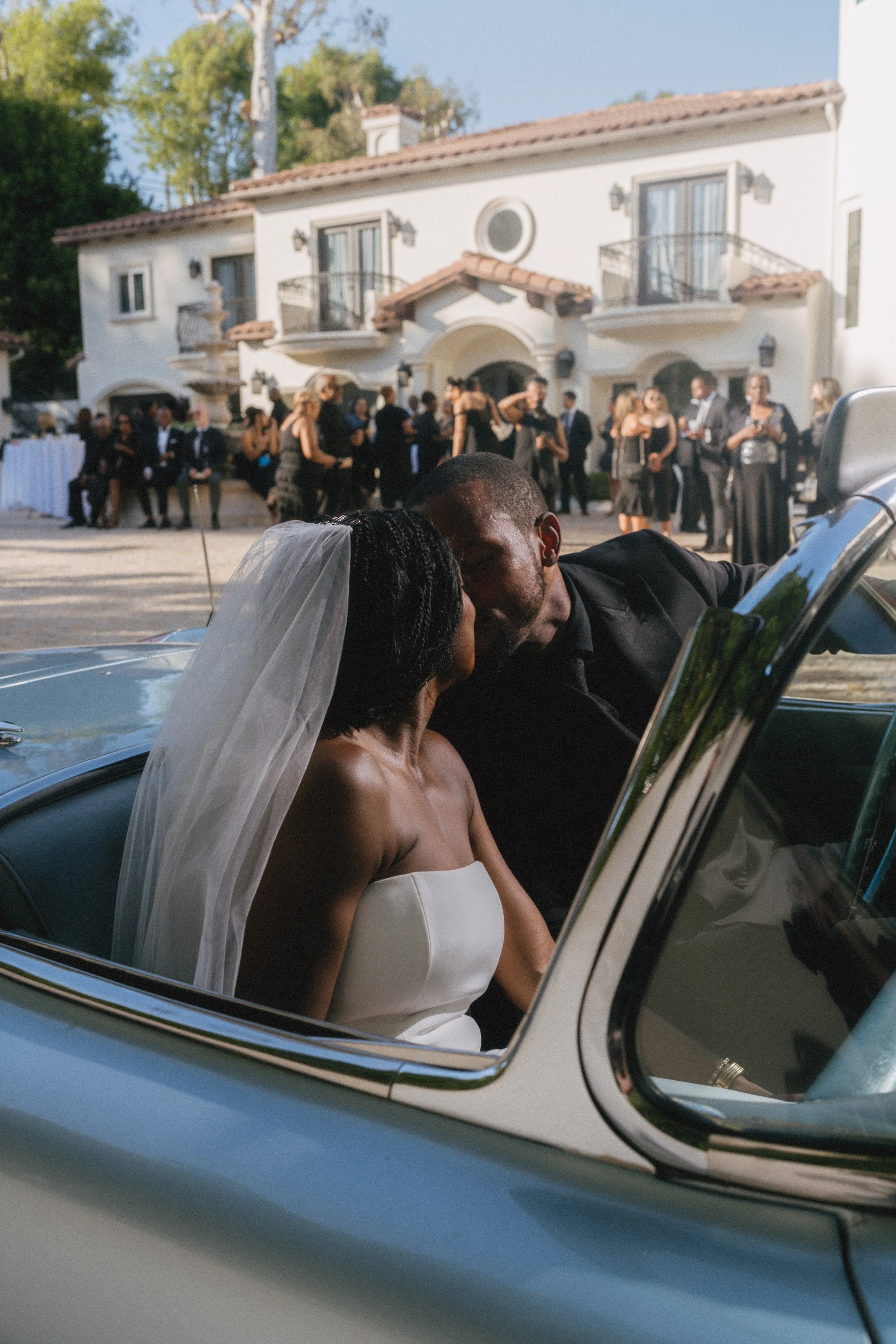 A newlywed couple sharing a kiss in a vintage car during their wedding celebration, with a large white house and guests gathered in the background.