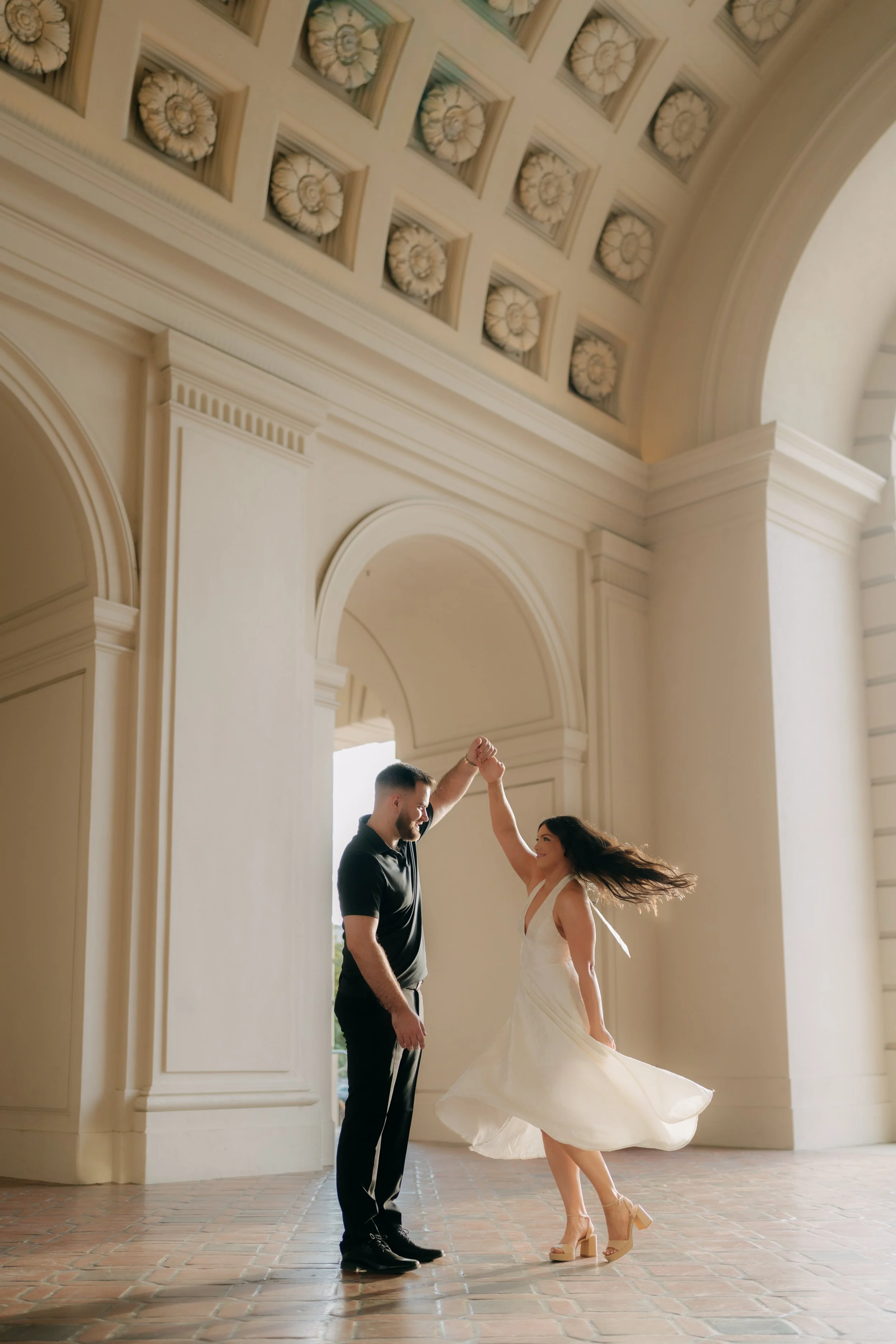 A couple dancing inside a spacious, classical-style building with large archways and decorative ceiling panels.