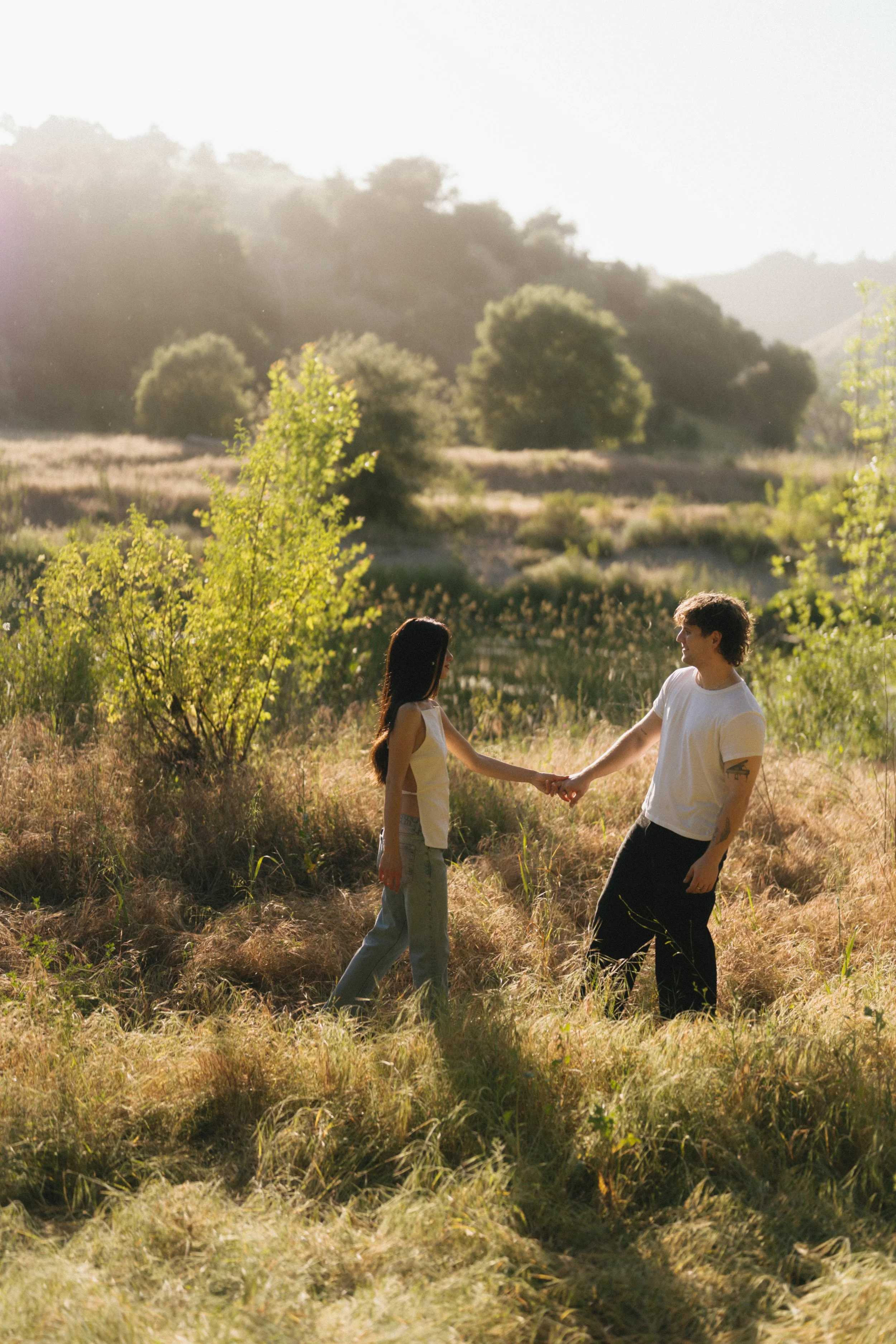 A young man and woman holding hands in a grassy field with trees and hills in the background, bathed in warm sunlight.