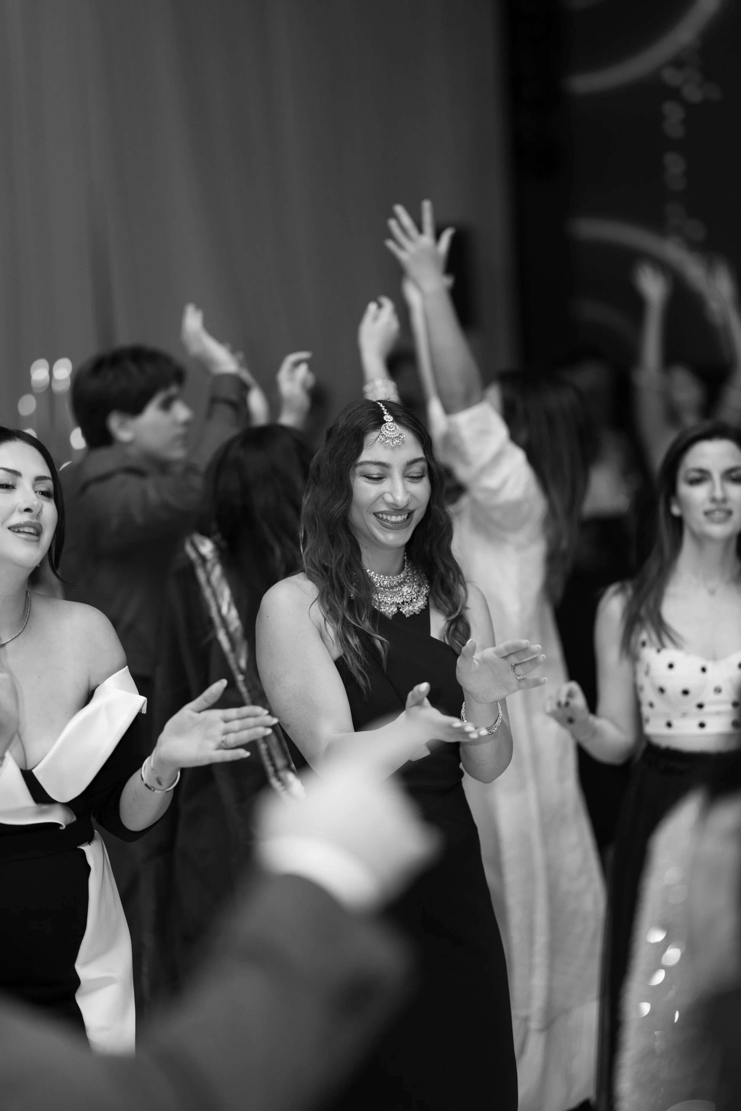 A women wearing traditional jewelry is clapping and smiling at a celebration or event, surrounded by other women and men raising their hands in the background.