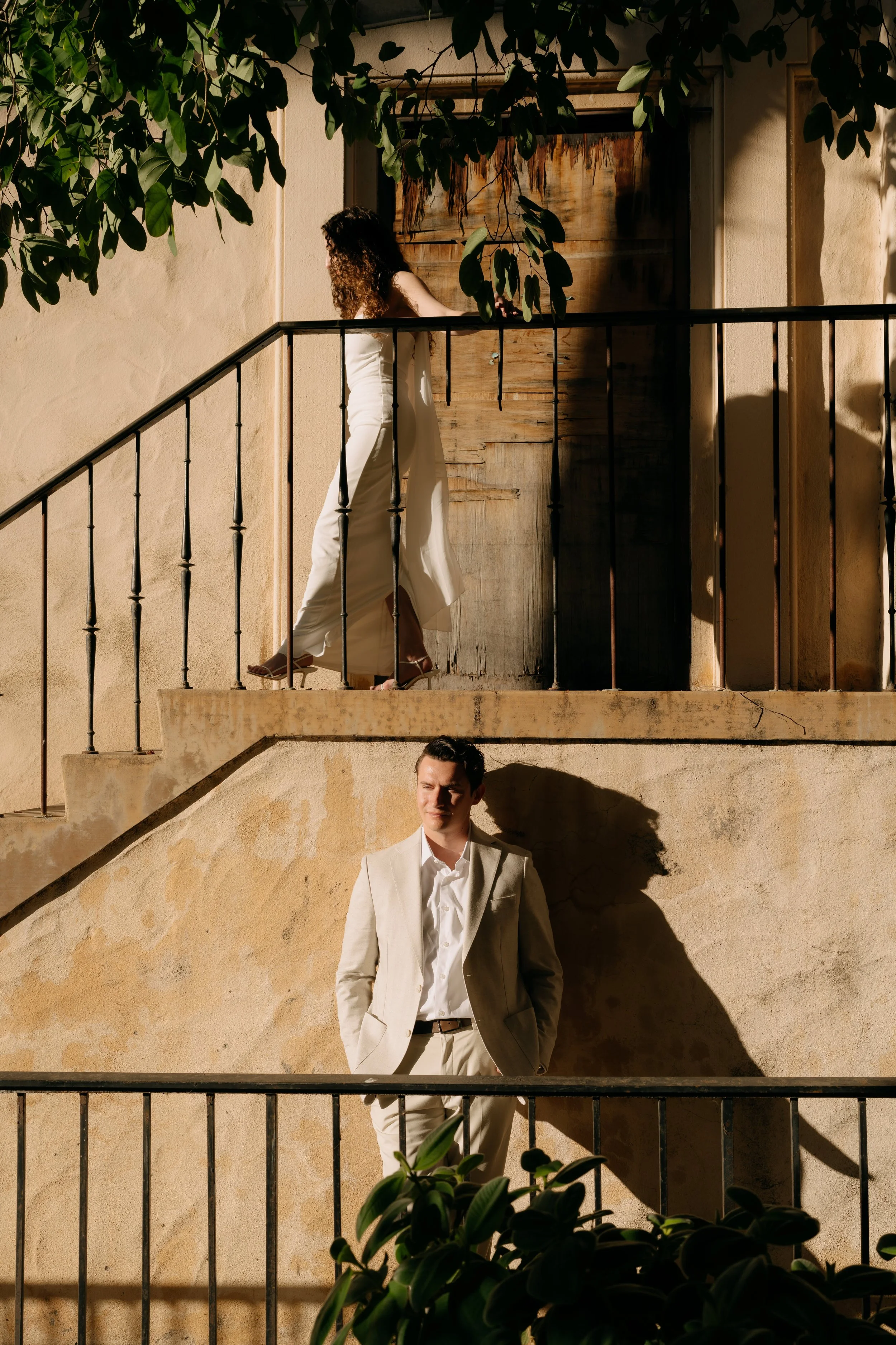 A woman with curly hair in a white outfit standing on a staircase balcony with greenery overhead. Below, a man in a beige suit stands against a beige wall with sunlight casting shadows.