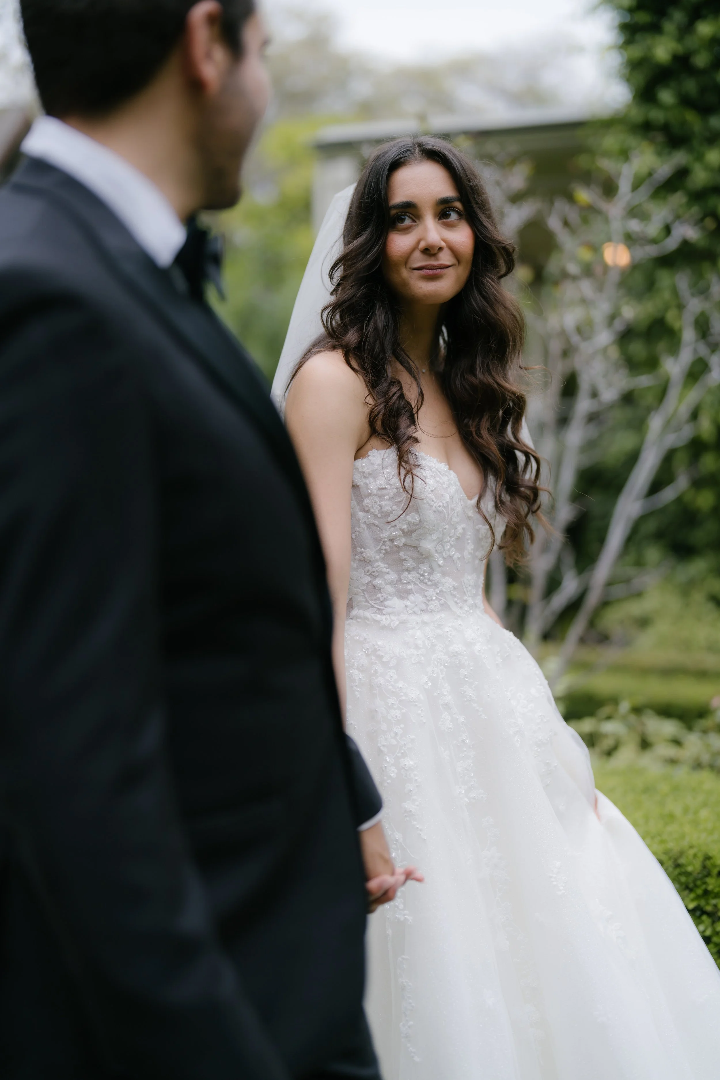 A bride and groom holding hands outdoors, with the bride looking at the groom as they stand in a garden or park setting.