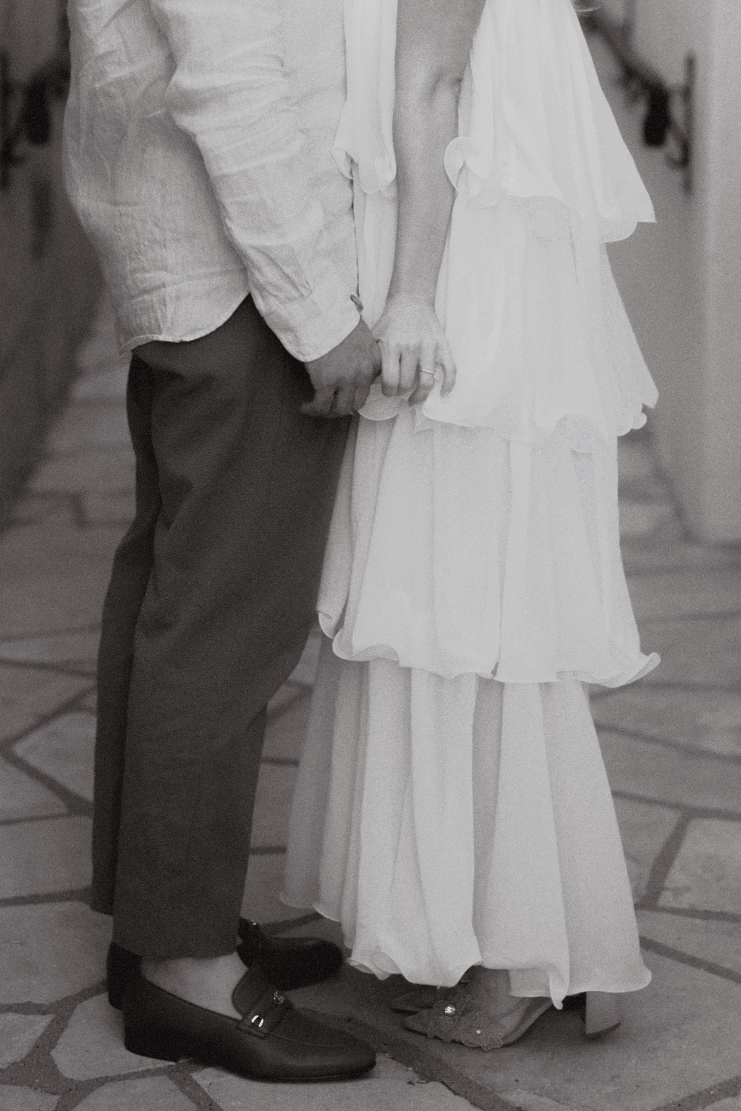 Close-up of a couple holding hands, with the woman wearing a white dress and the man wearing dark pants and shoes, on a tiled floor.