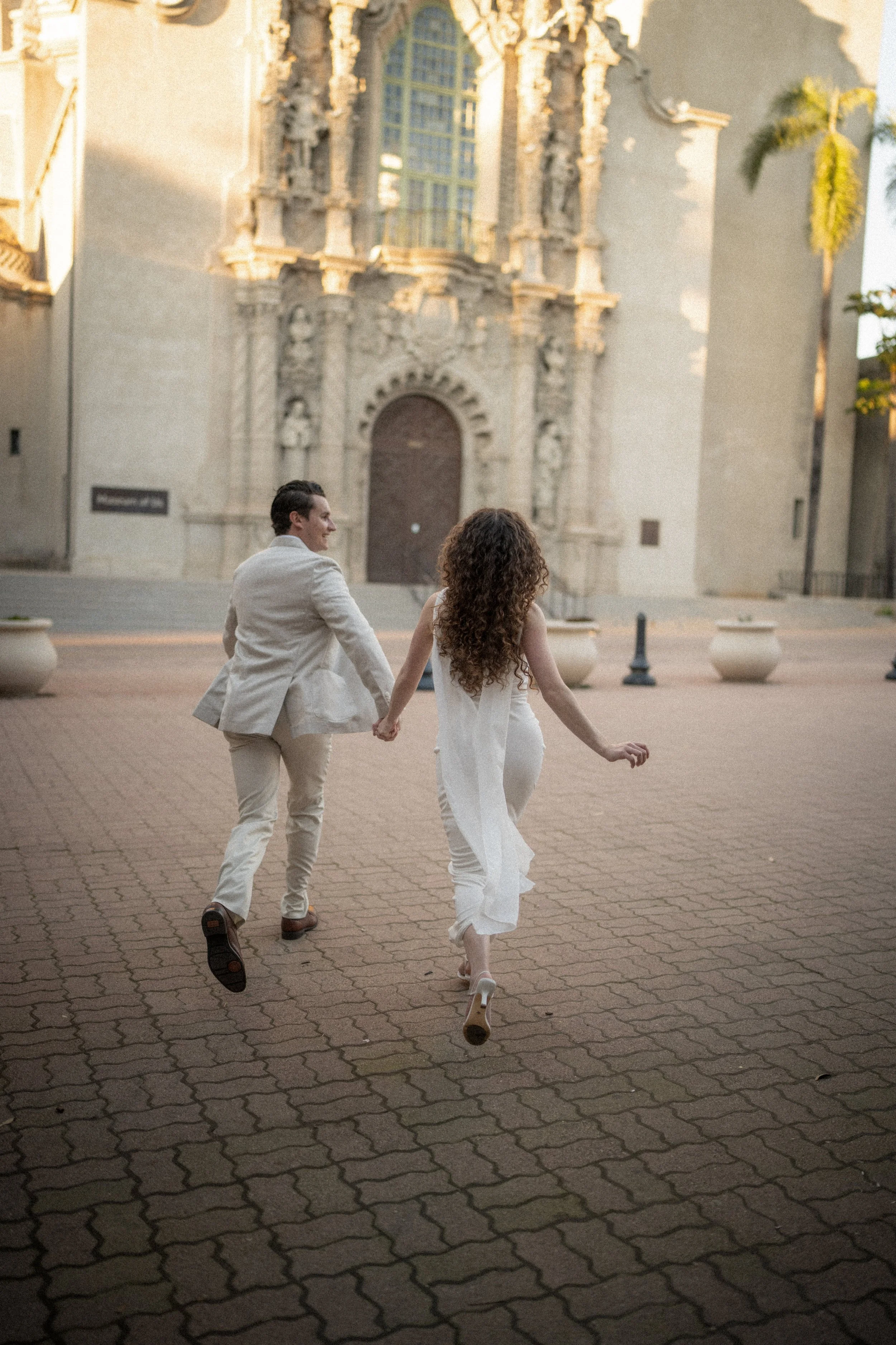 A couple holding hands and running on a brick sidewalk in front of an ornate building with a large window and decorative stonework, during sunset.