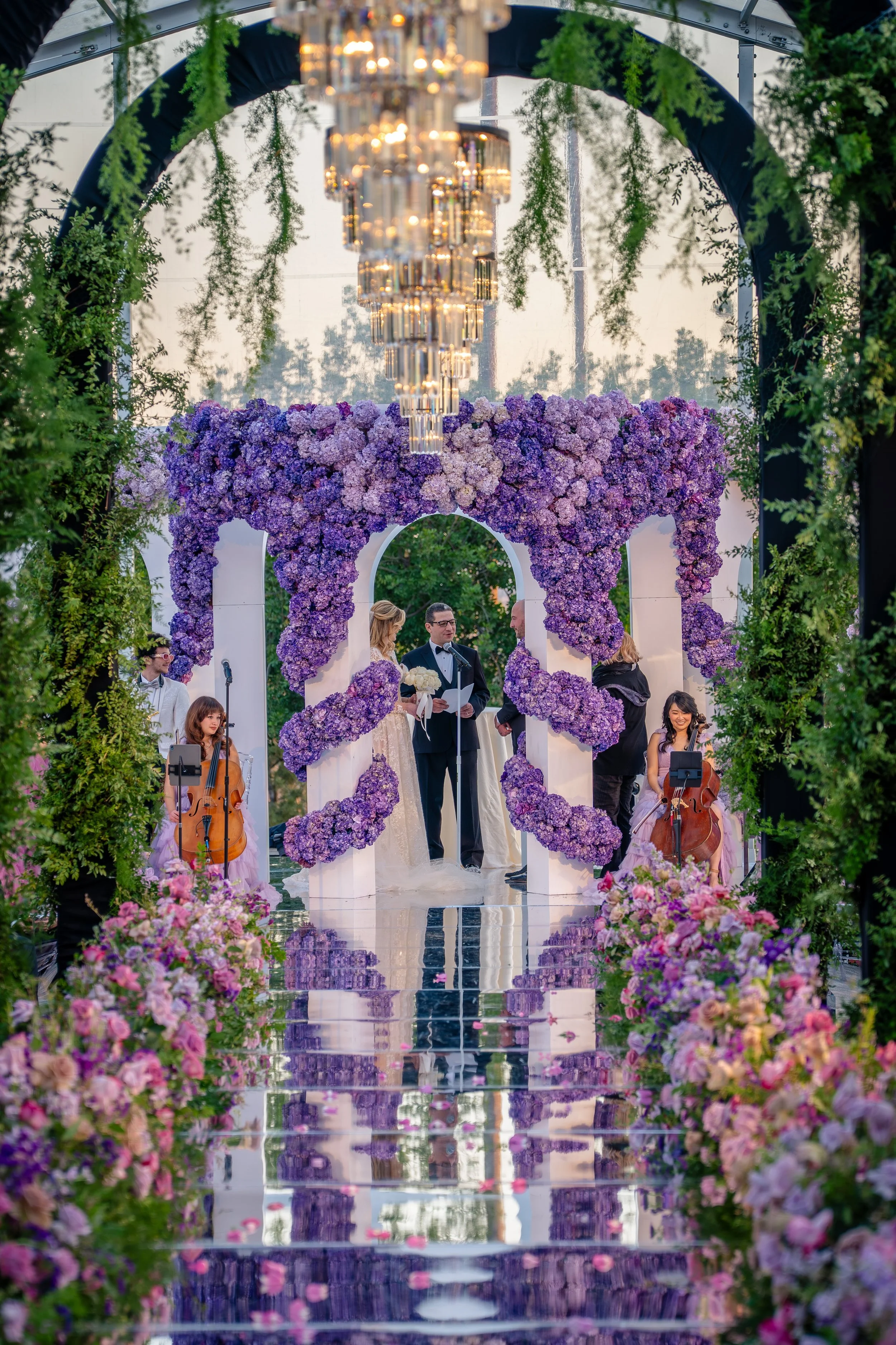 A wedding ceremony taking place outdoors under a floral arch decorated with purple and pink flowers, with string musicians playing and a chandelier hanging above.