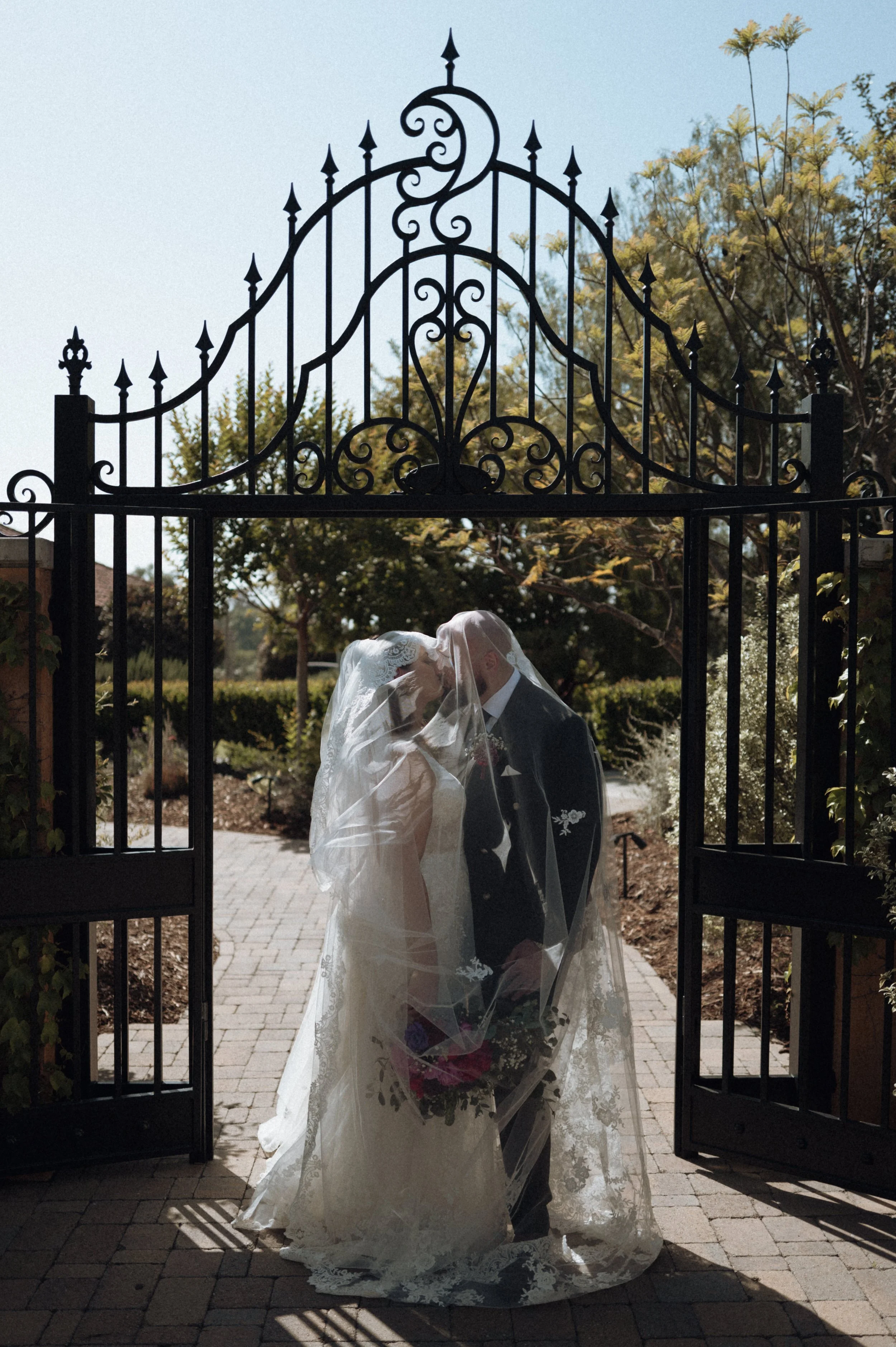 A bride and groom share a kiss under a black wrought-iron gate during their wedding, with trees and a clear sky in the background.