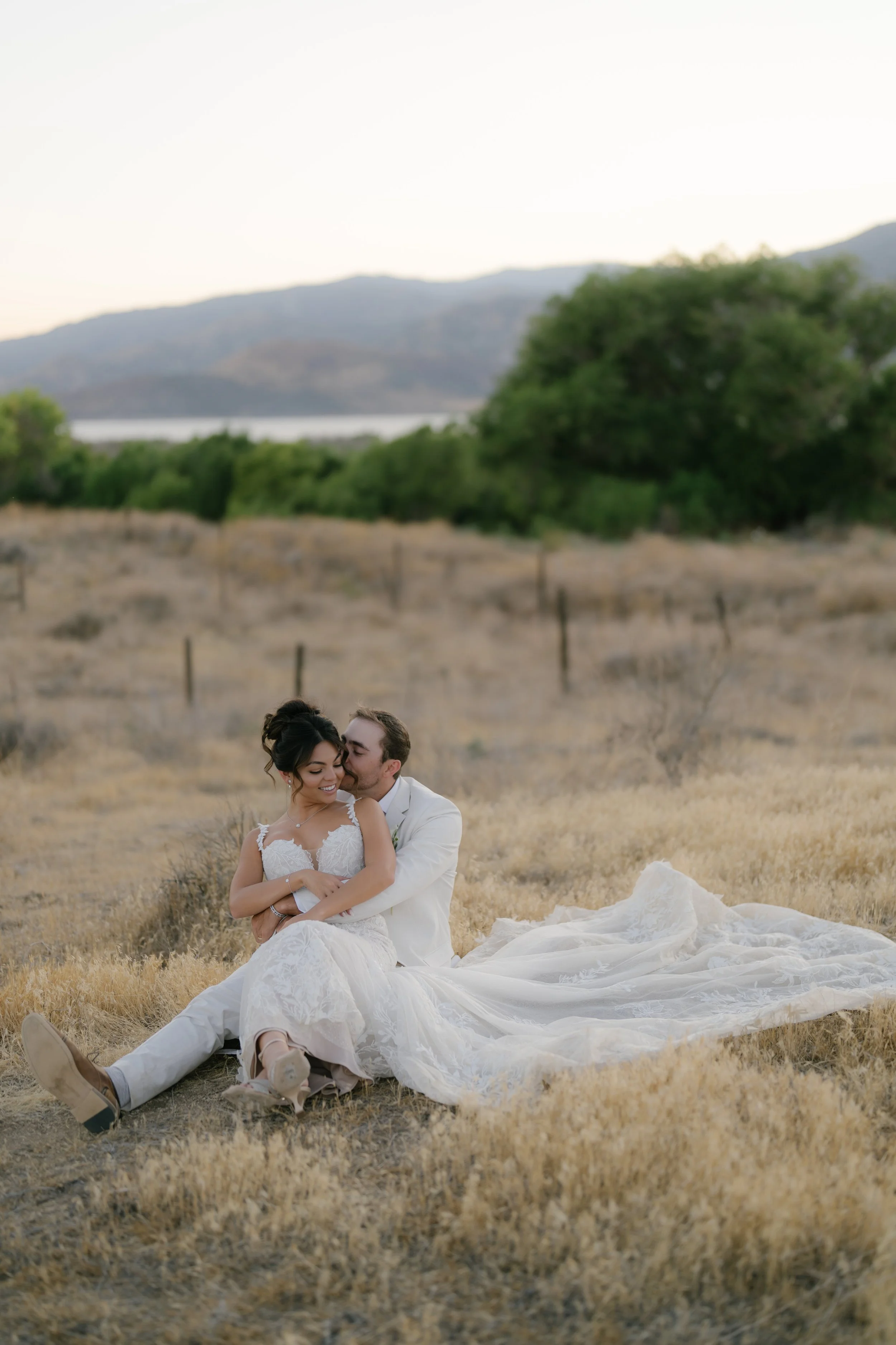 A newlywed couple sitting on dry grass in a field with mountains and trees in the background. They are dressed in wedding attire, with the bride wearing a white lace gown and the groom in a white suit. The groom is embracing the bride from behind, an