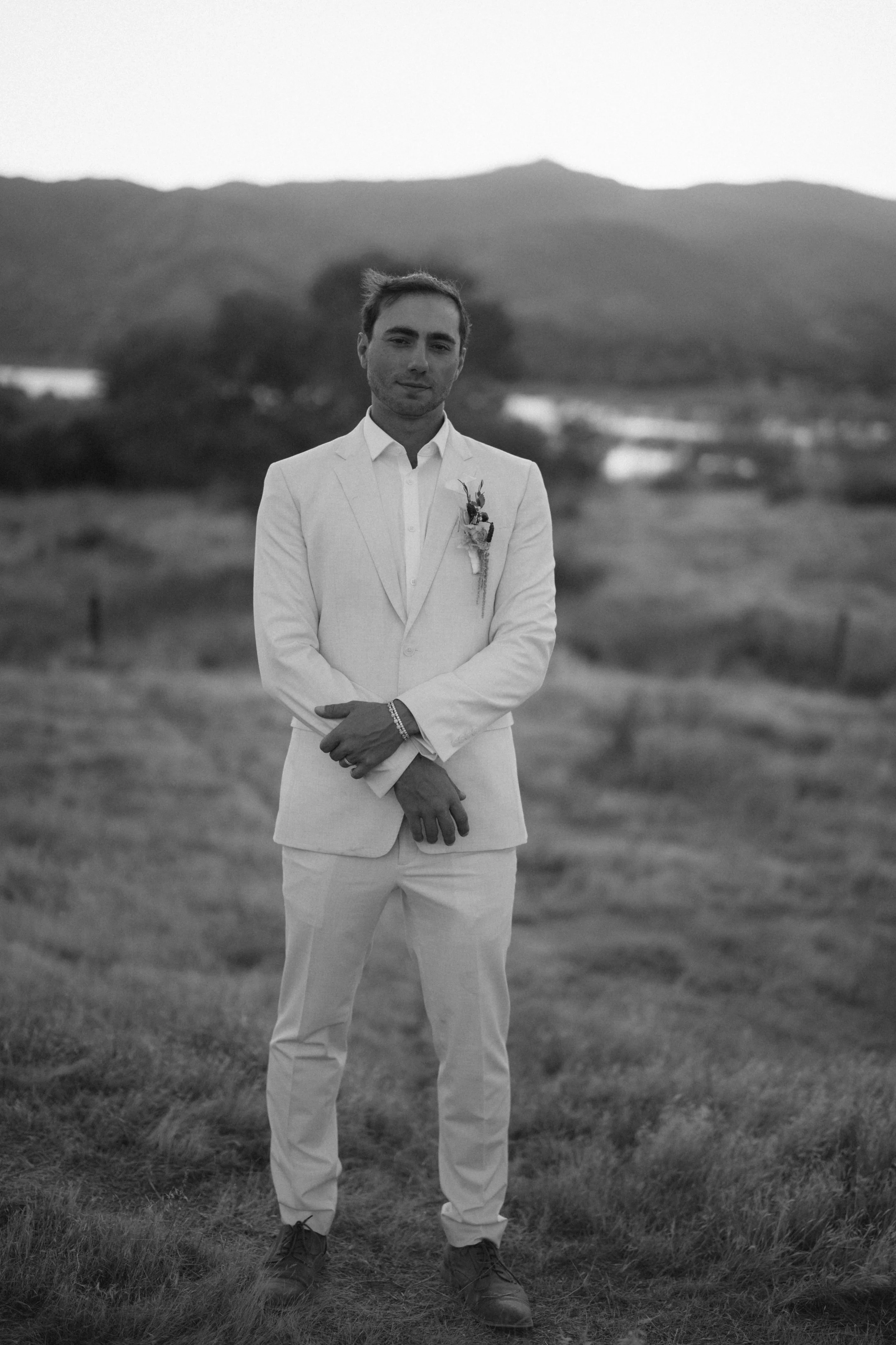 A young man in a light-colored suit and tie standing outdoors on grass with mountains in the background in black and white.