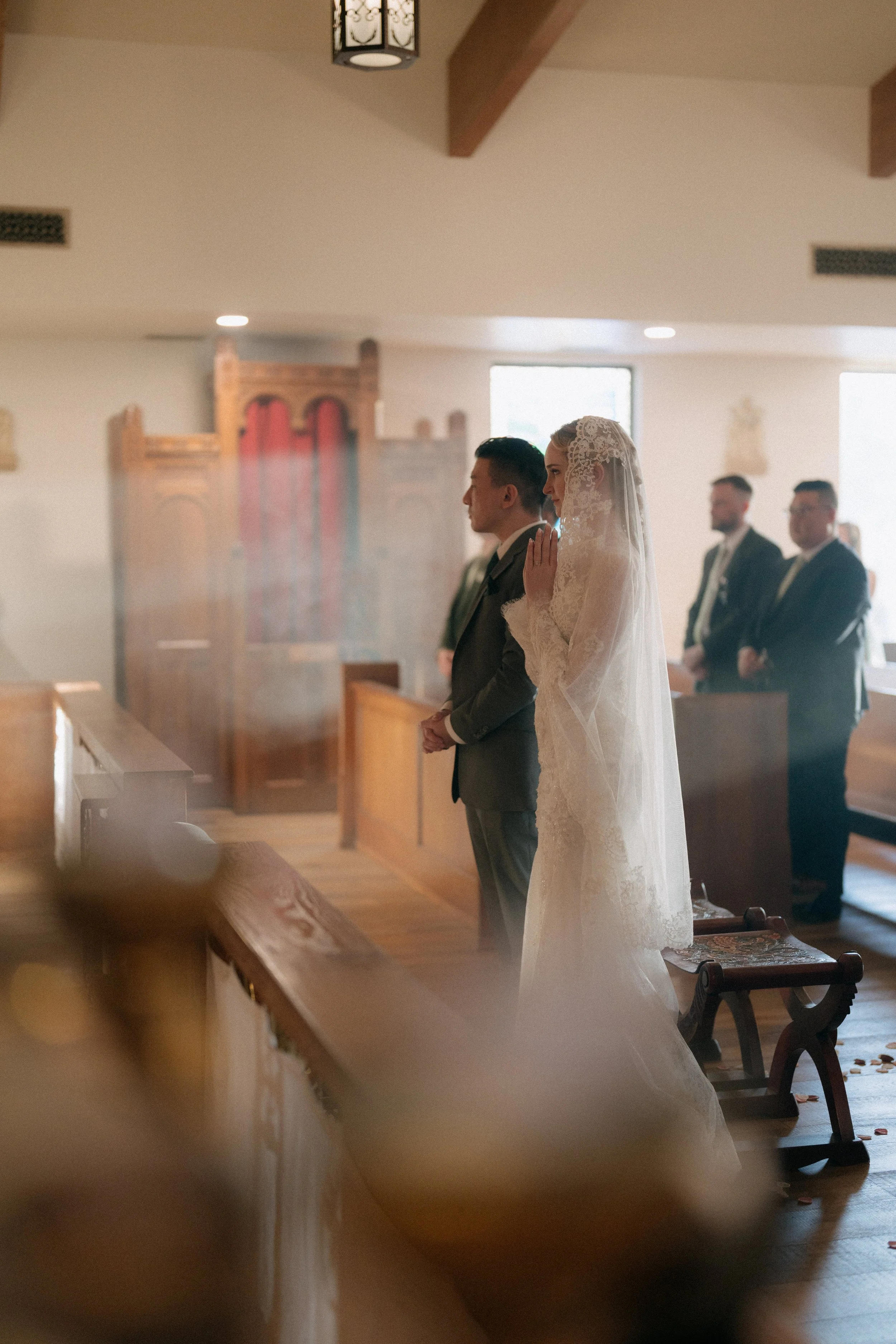 A bride in a lace wedding dress and veil stands with her hands pressed together in prayer or reflection during a wedding ceremony, with groomsmen in suits in the background inside a church.