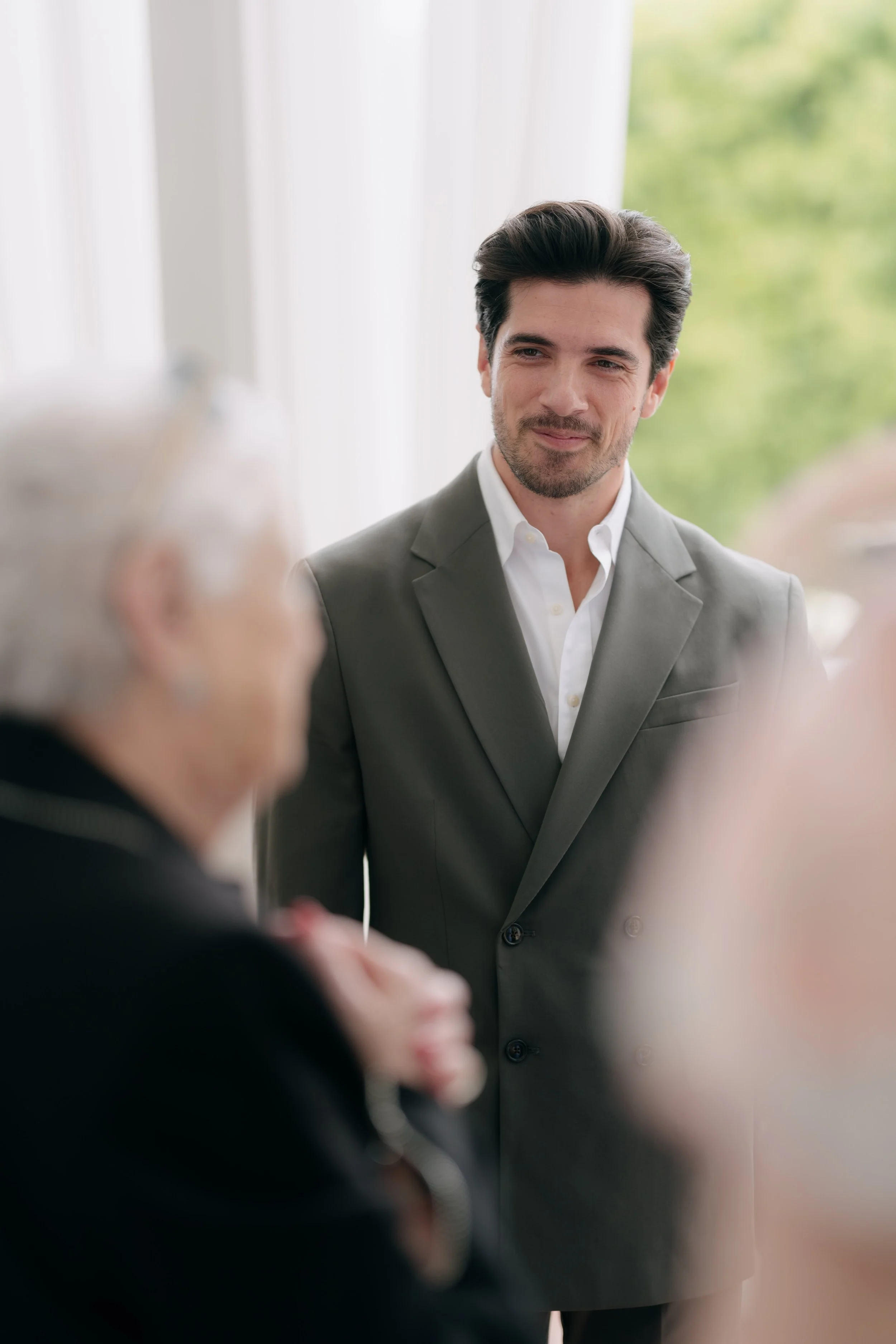 A man with dark hair and a beard, wearing a light gray suit and white shirt, is smiling and looking at an older man with gray hair during a conversation.