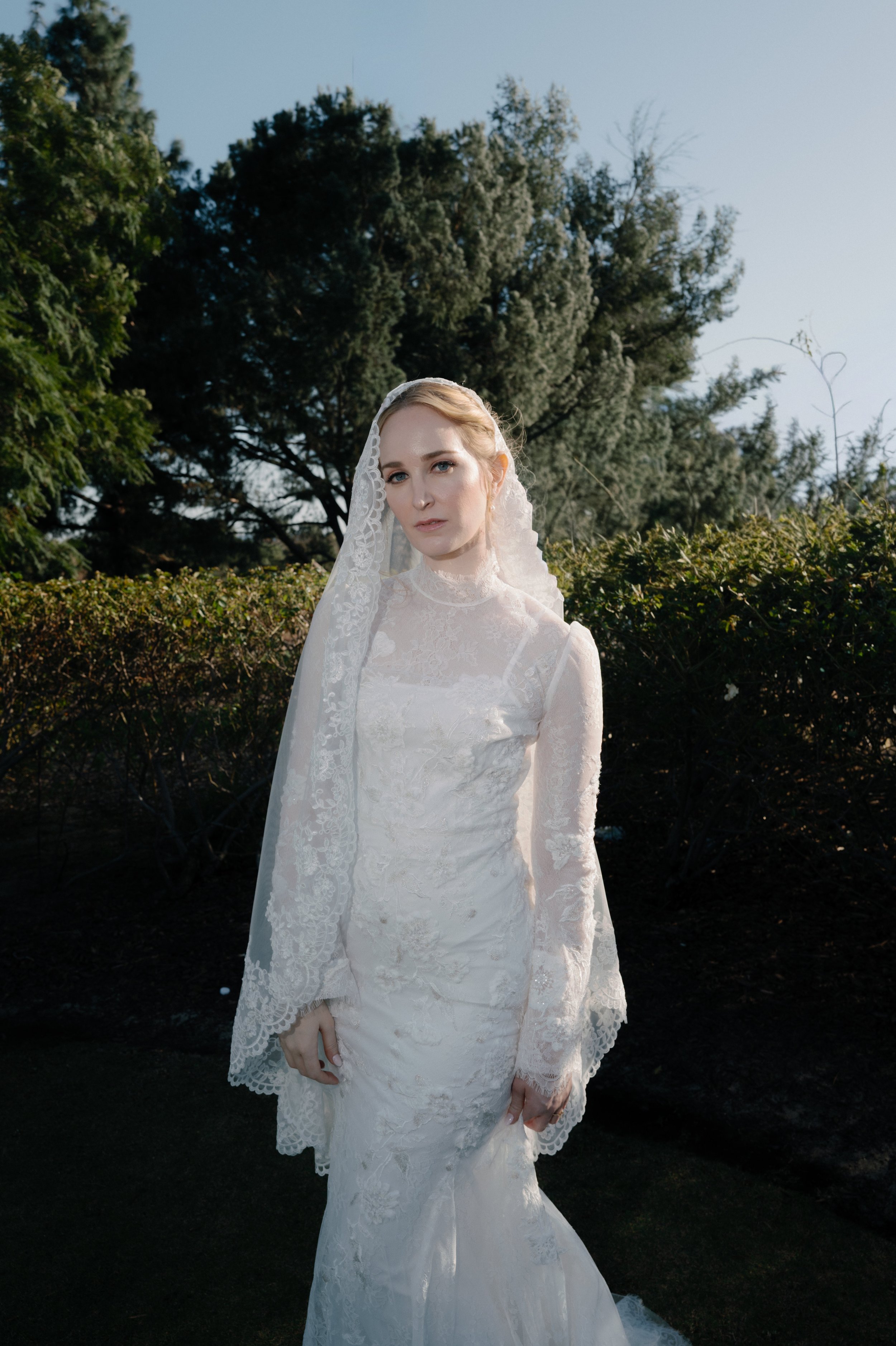 A woman dressed in a vintage lace wedding gown with a veil, standing outdoors in front of lush green bushes and trees on a sunny day.
