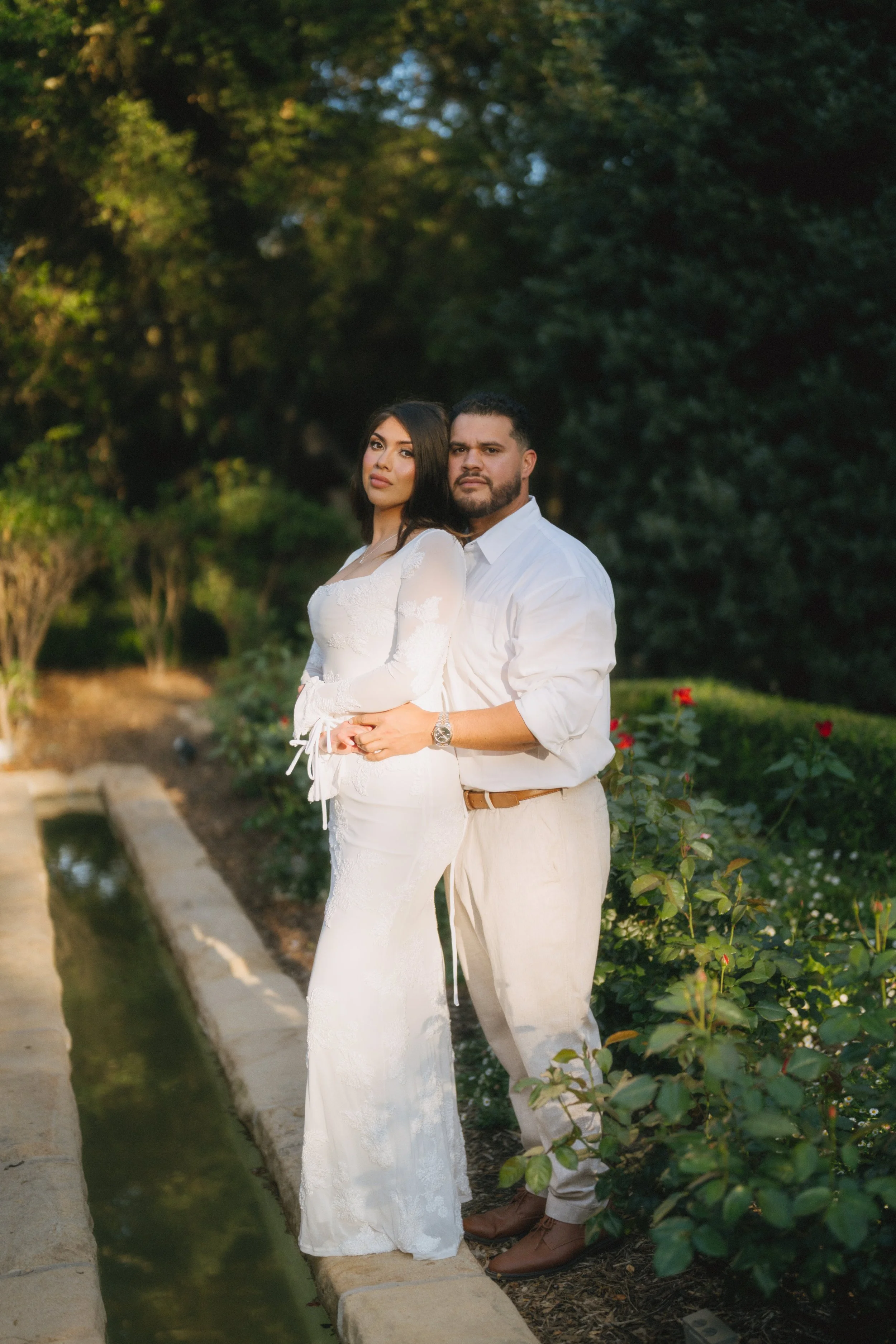 A couple standing together outdoors near a small water feature, with trees and bushes in the background during daytime.