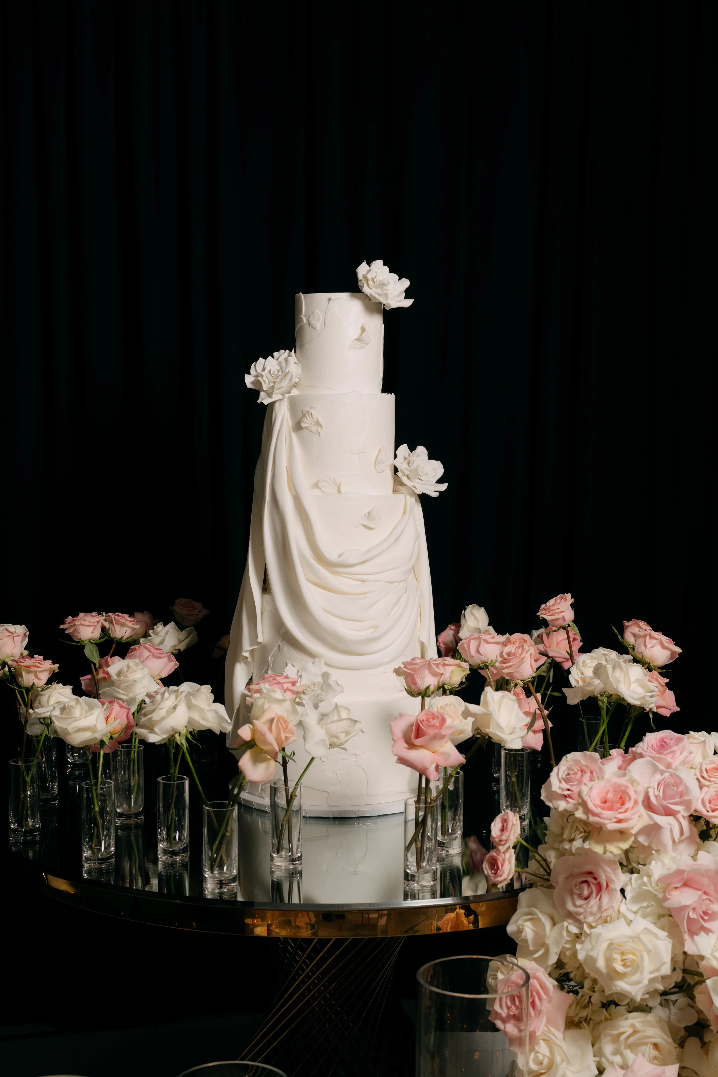 A three-tiered white wedding cake decorated with white flowers and leaves, surrounded by pink and white roses in glass vases on a black table with a dark background.
