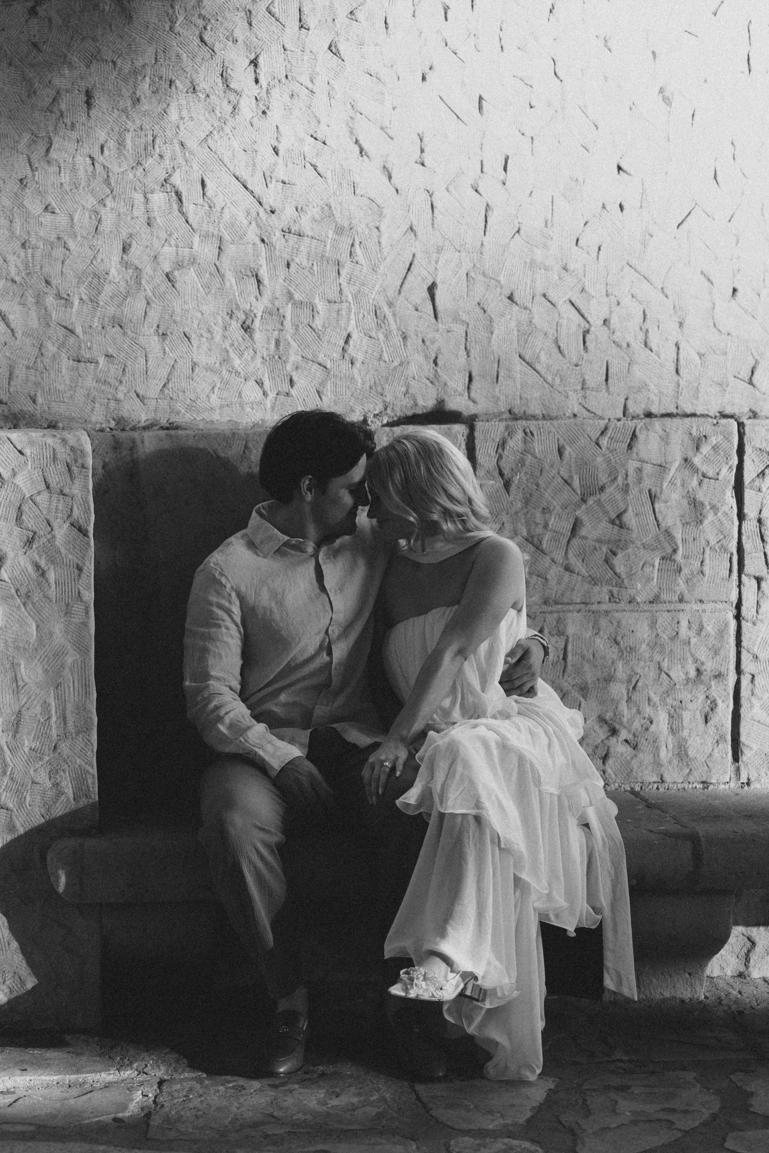 A man and a woman share an intimate moment on a bench against a textured stone wall, with the woman wearing a flowing dress and the man dressed in a shirt and trousers.