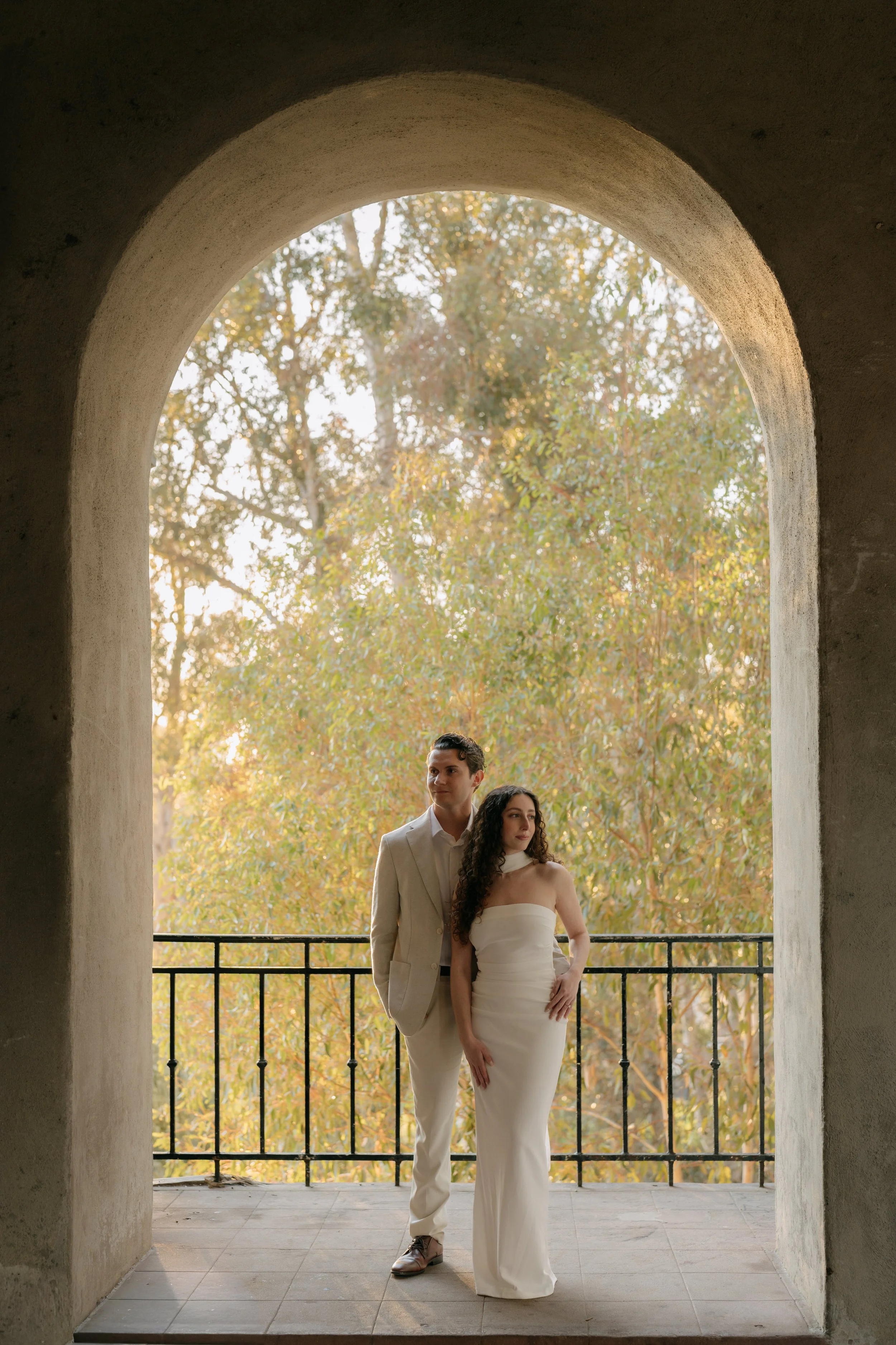 A couple standing on a balcony framed by an arched stone opening with trees in the background. The woman wears a strapless white dress, and the man wears a light-colored suit.