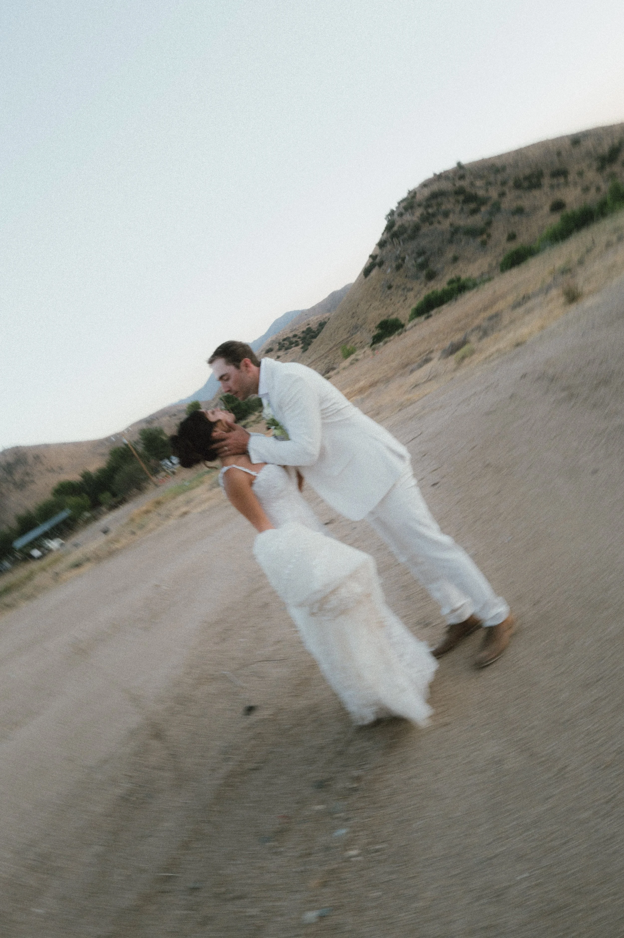 A couple in wedding attire dancing together outdoors on a dirt path with hills and sparse vegetation in the background.