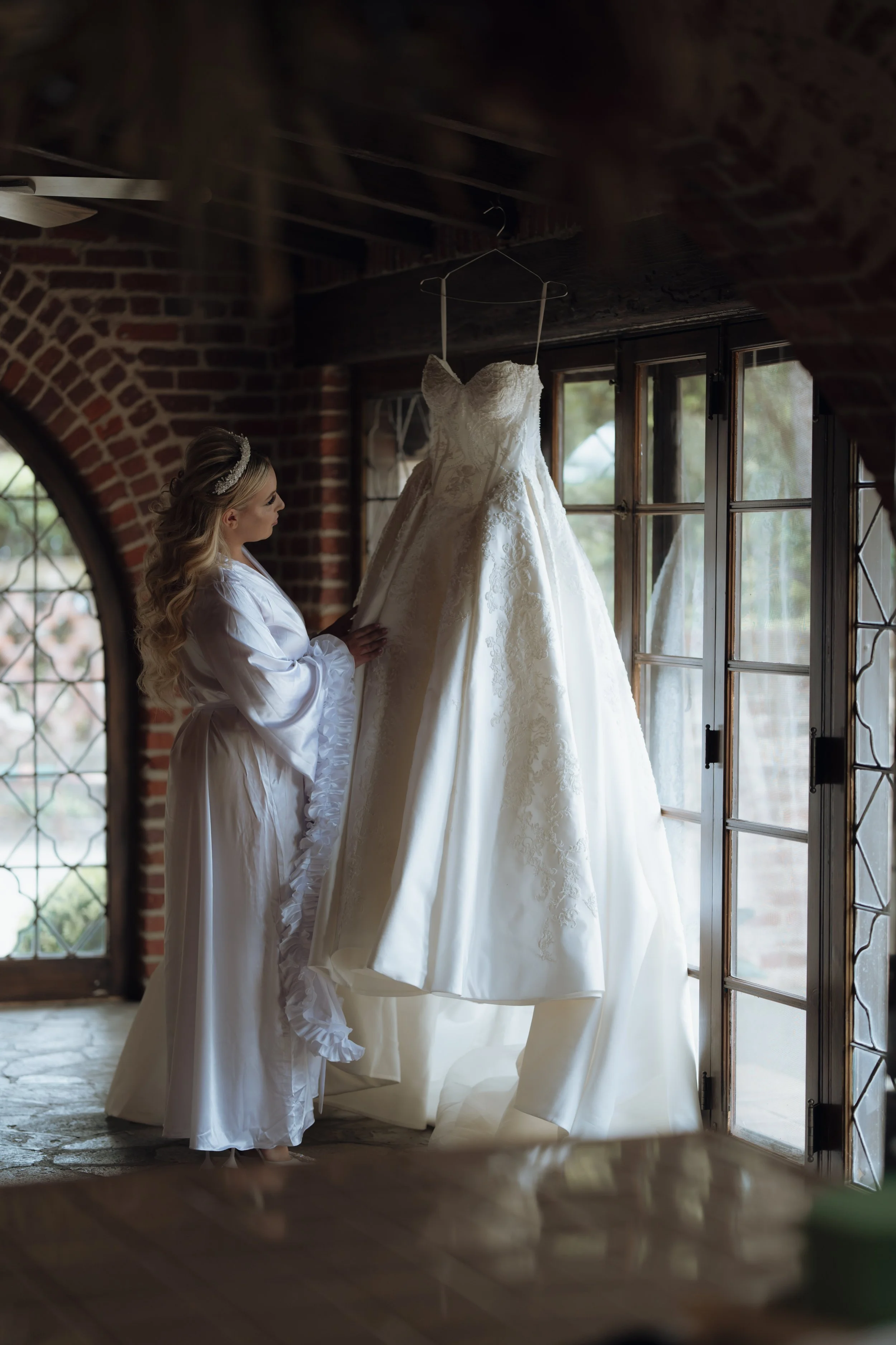 A woman with long blonde hair wearing a white satin robe with ruffled sleeves, looking at a white wedding dress hanging in front of her by a windowsill in a rustic brick room.