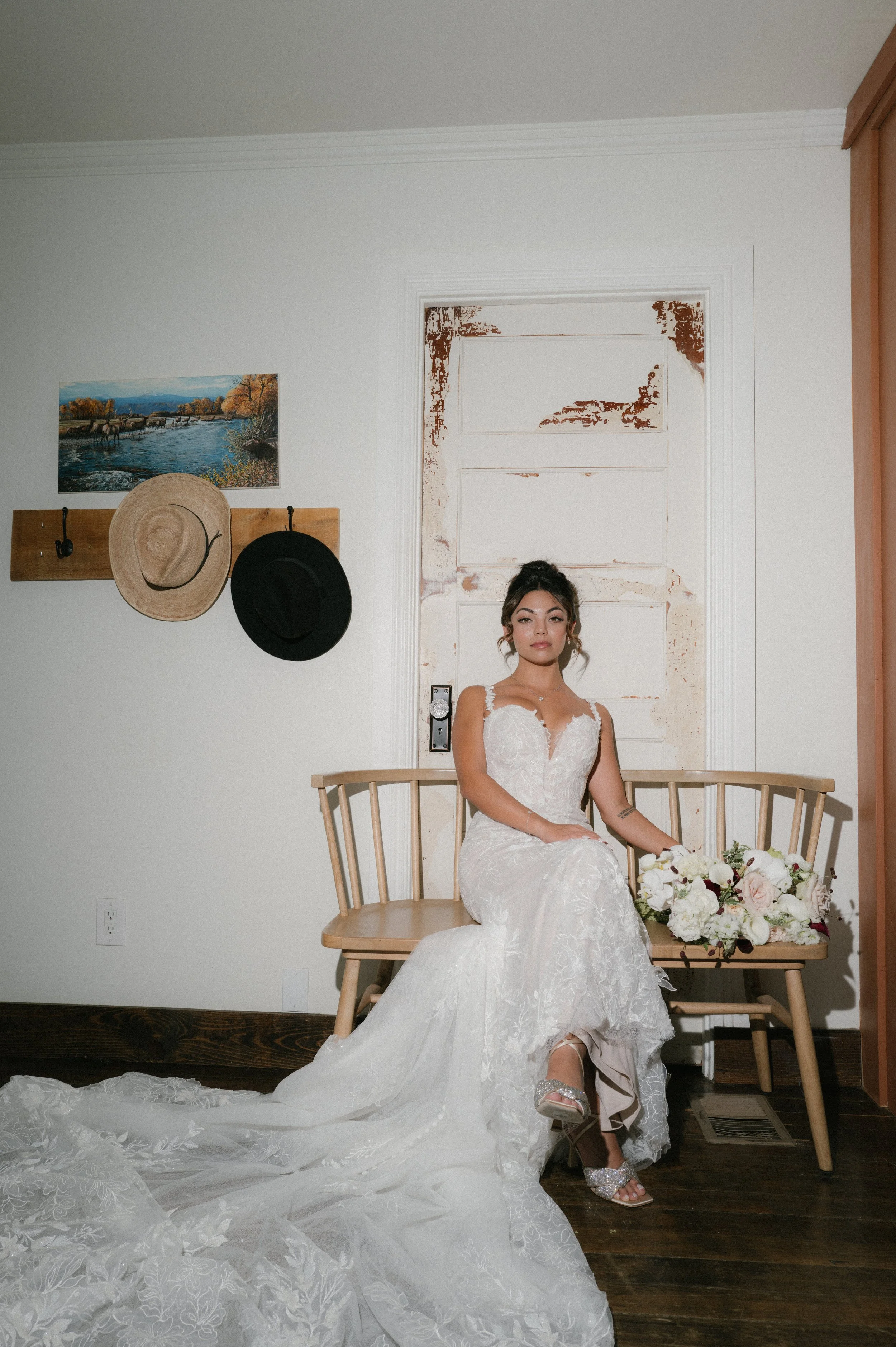 A woman in a white wedding dress sitting on a wooden bench in a room with white walls. There are two hats hanging on a wooden coat rack on the wall and a painting of a river scene with trees and mountains. The woman has a bouquet of white and pink fl