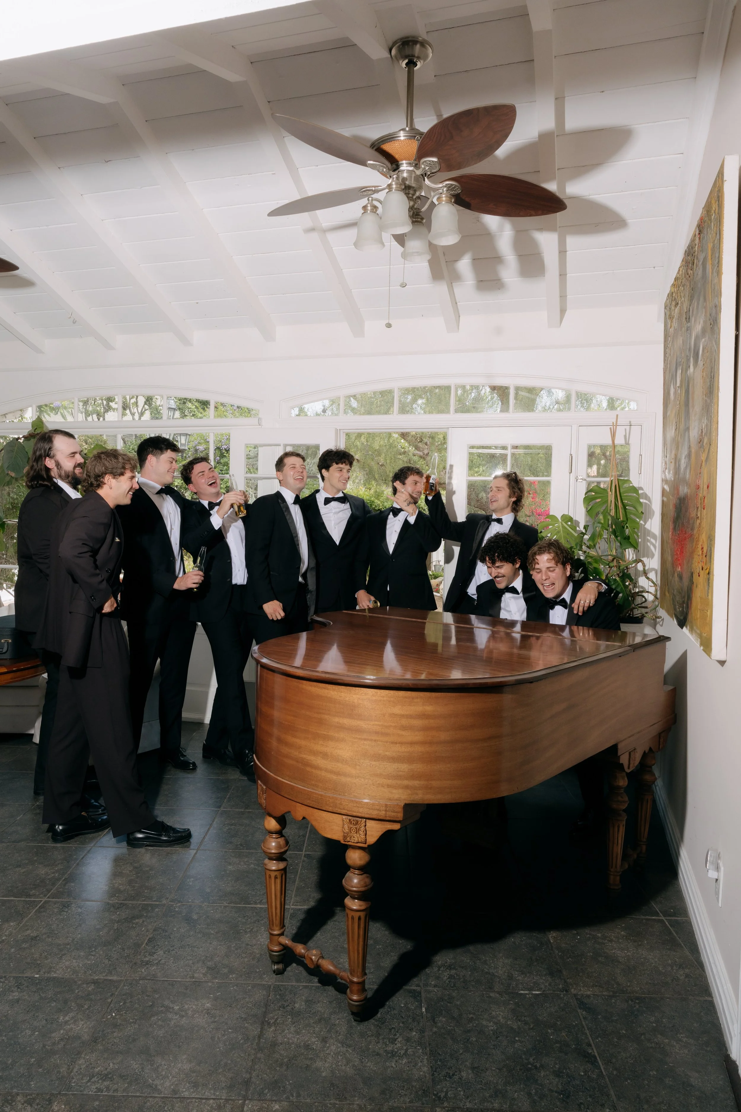 A group of men dressed in tuxedos celebrating around a grand piano in a bright room with large windows, plants, and ceiling fans.