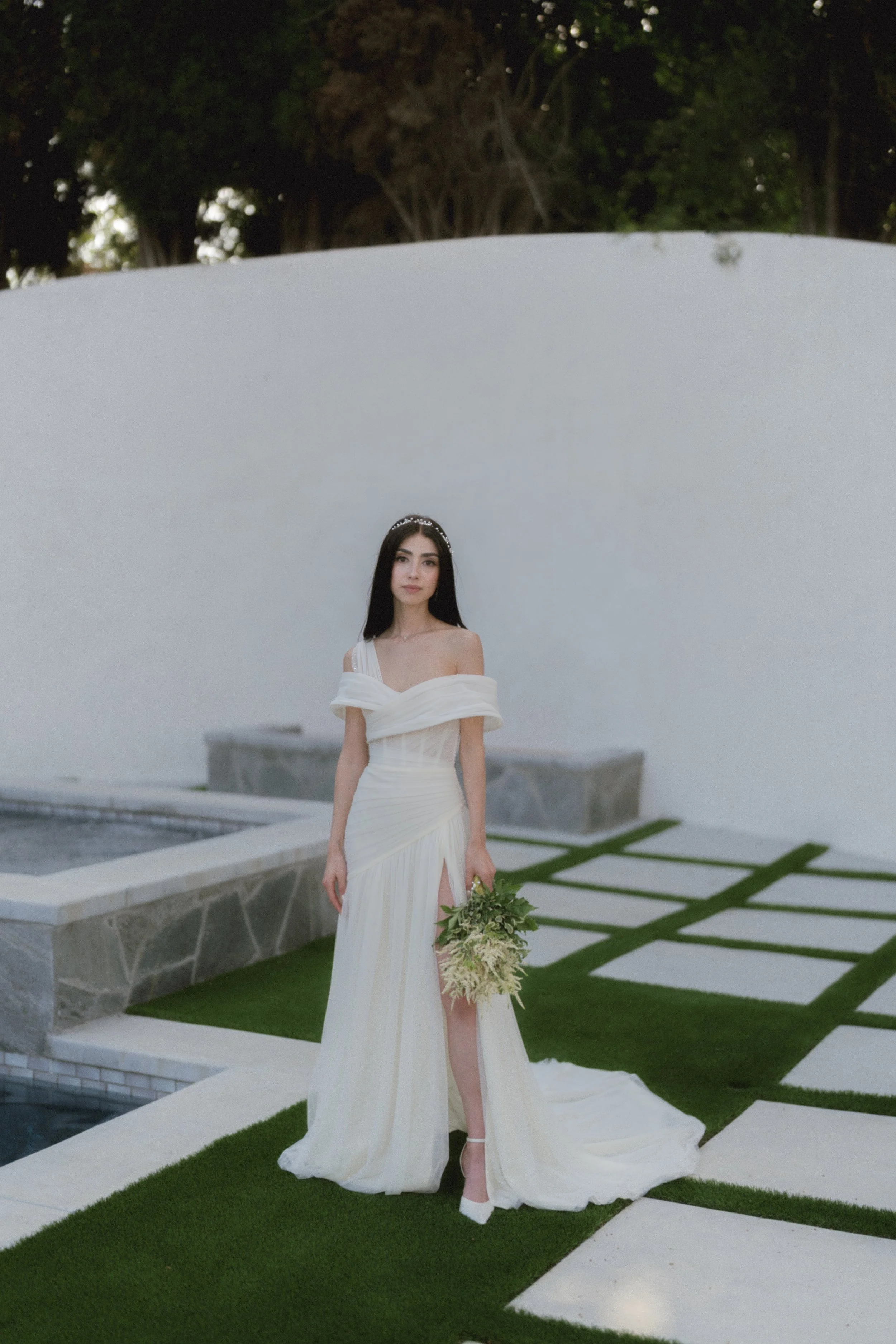 A woman in a white wedding dress with a high slit, veil, and tiara holding a bouquet of white and green flowers standing outdoors near a modern white wall and geometric garden design.