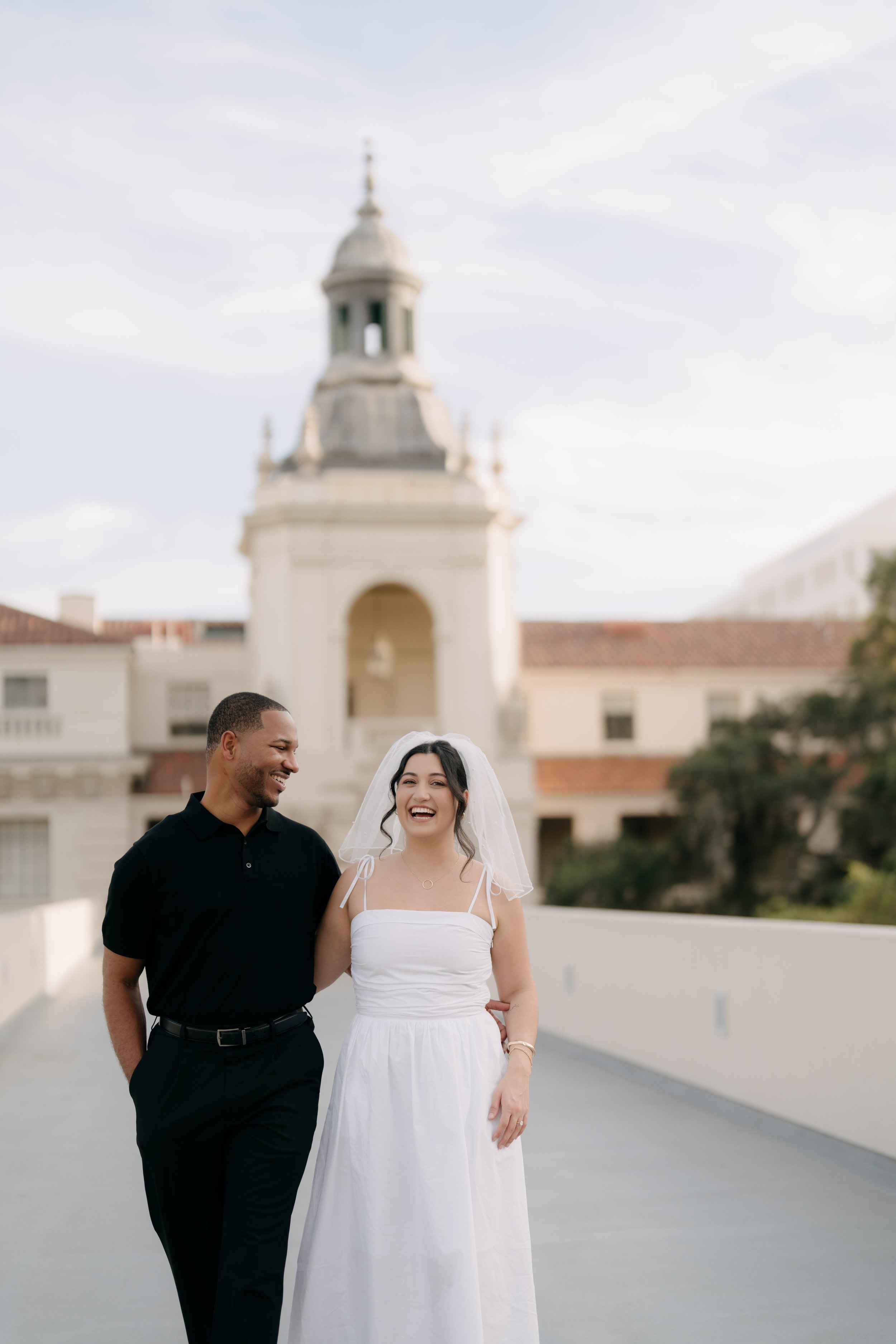 A smiling couple, a man in a black shirt and a woman in a white dress with a veil, standing together outdoors near a building with a clock tower, on a cloudy day.