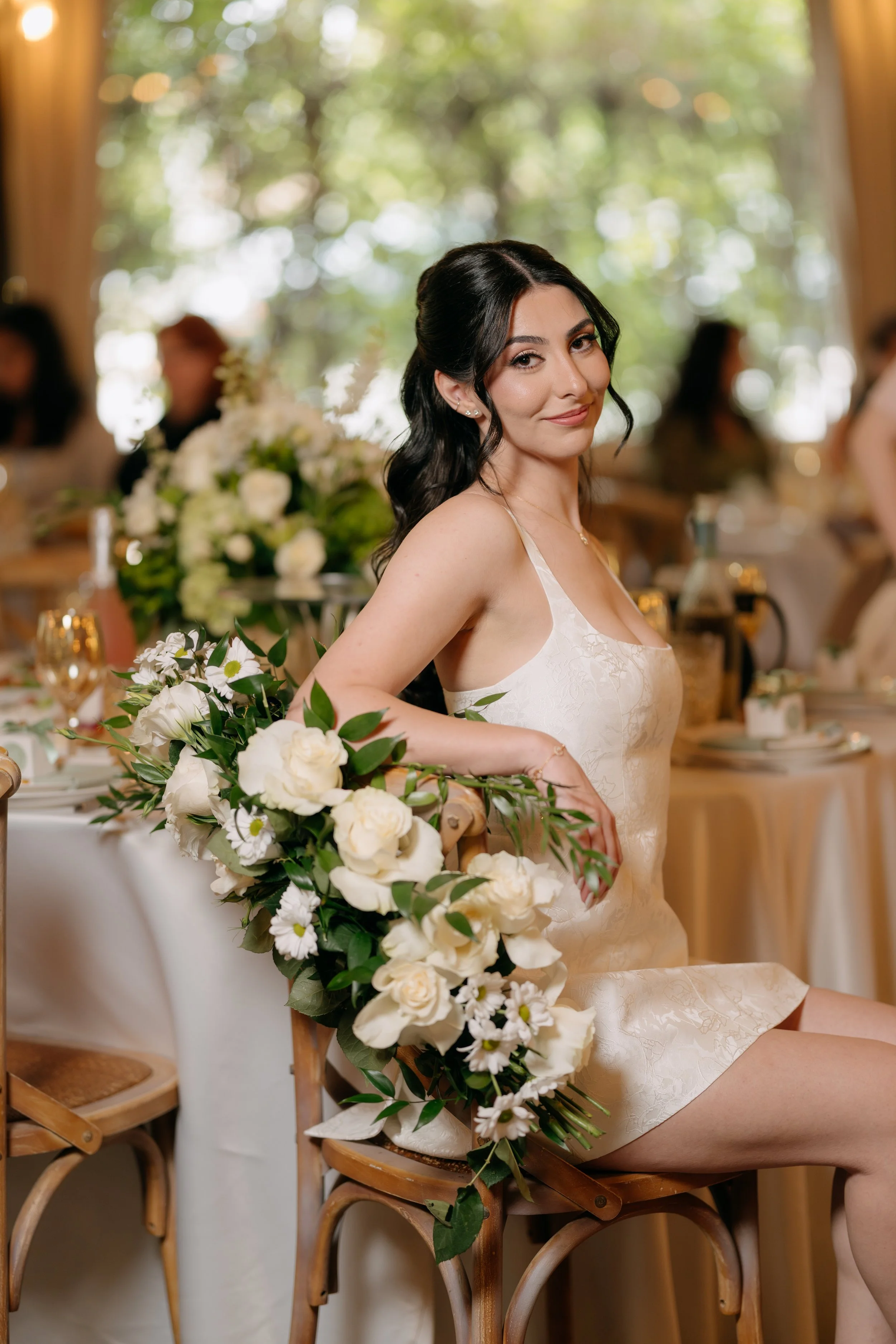 A bride sitting at a wedding reception with a floral arrangement in front of her, smiling and looking at the camera.